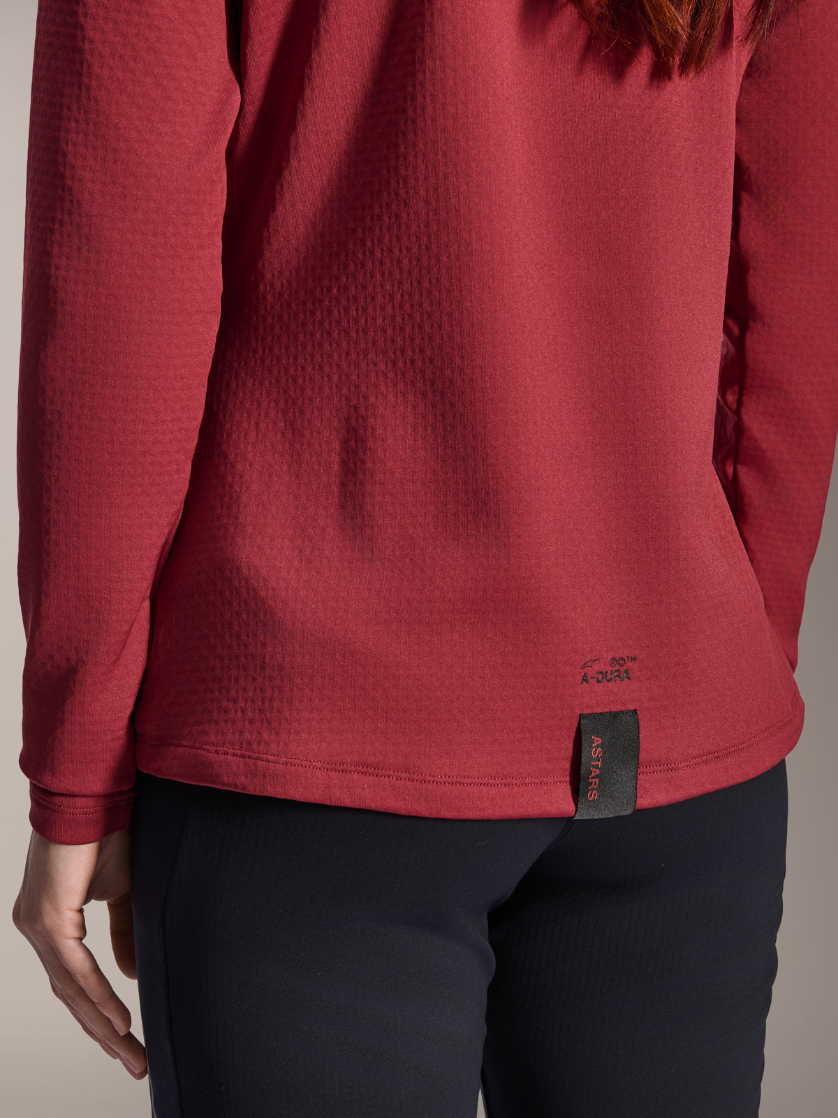 Camisola Térmica Feminina Alpinestars Stella A-Dura, Borgonha, vista de pormenor da bainha traseira com a marca A-Dura e a etiqueta tecida ASTARS, tecido polar isolante texturizado, vestuário de BTT para mulher