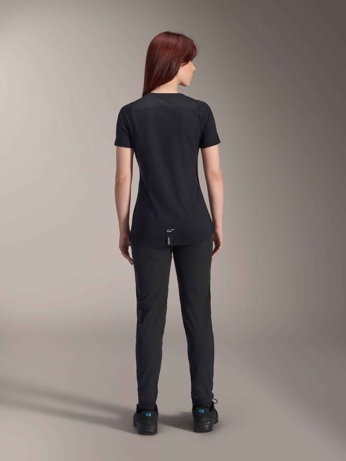 Camisola de manga curta feminina Alpinestars Stella A-Dura Dri Switch, preta, vista traseira numa modelo, com cauda mais longa e painéis de malha técnica, combinada com calças pretas, equipamento de ciclismo MTB de alta performance