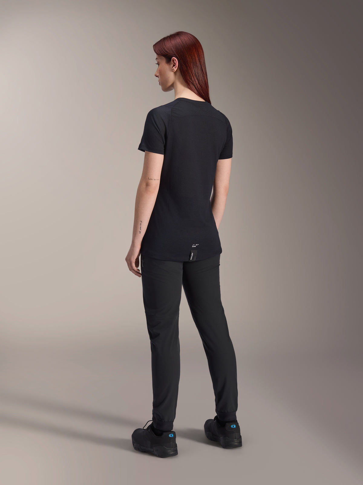 Modelo feminina de cabelo ruivo a usar a camisola de manga curta Alpinestars Stella A-Dura Dri Switch em preto, vista traseira, combinada com calças e sapatos de BTT pretos, realçando o ajuste técnico e a parte traseira mais comprida para maior cobertura em ciclismo.