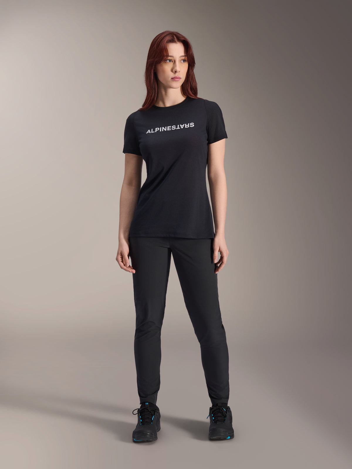 Modelo a usar a camisola de manga curta Alpinestars Stella A-Dura Dri Switch para mulher em preto, com logótipo branco no peito, calças técnicas pretas e sapatilhas pretas, fotografia de estúdio de corpo inteiro