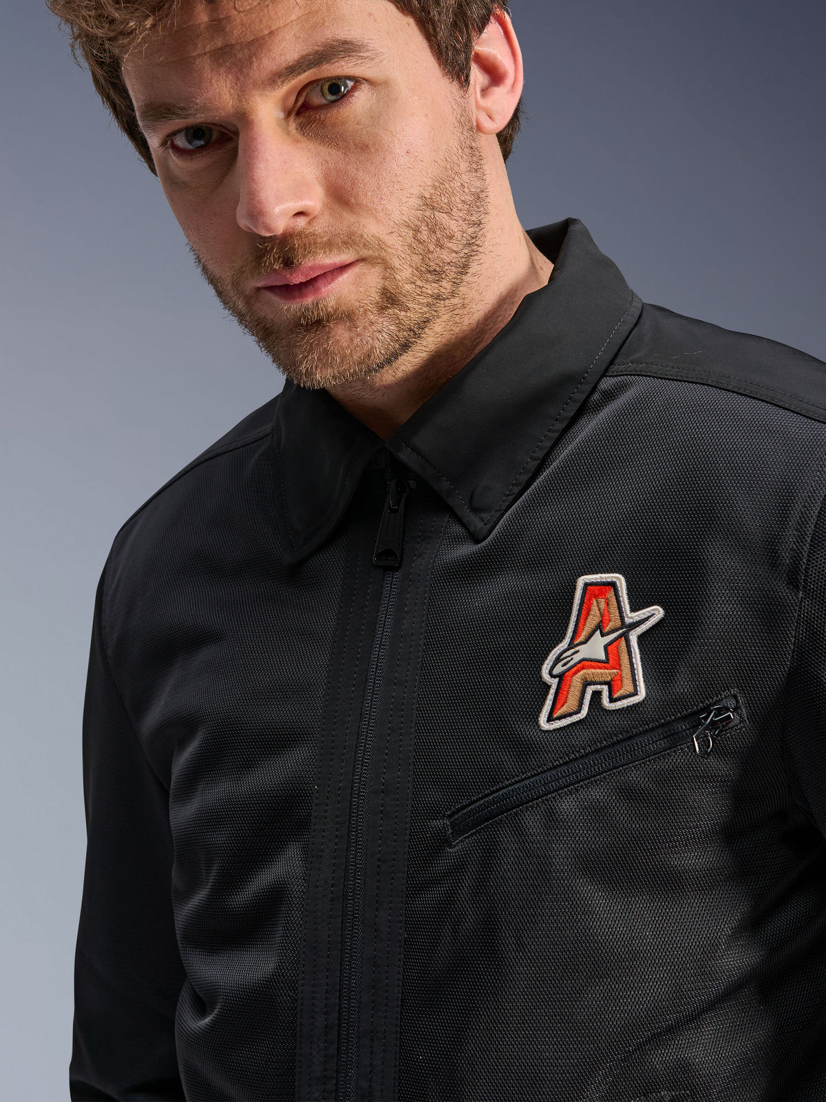 Casaco Alpinestars Ignite Air, preto, com gola de camisa tradicional e construção em malha respirável, realçado por um emblema com o logótipo 'A' em vermelho e dourado no peito, vestido por um modelo masculino, vestuário de motociclismo urbano
