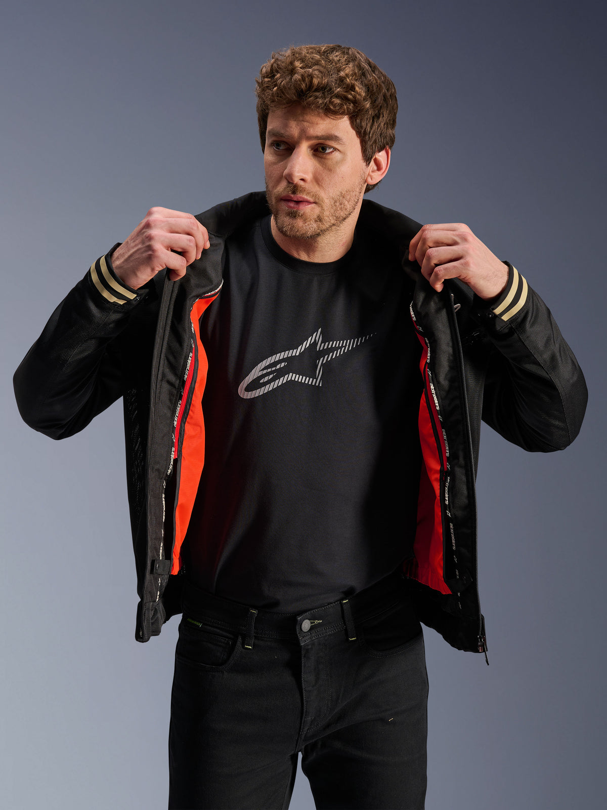 Homem a usar o Casaco Alpinestars Ignite Air, preto, com corpo em malha e forro corta-vento amovível em cor de laranja vibrante, t-shirt preta com o logótipo Astars e calças pretas, num estilo de motociclismo urbano contemporâneo