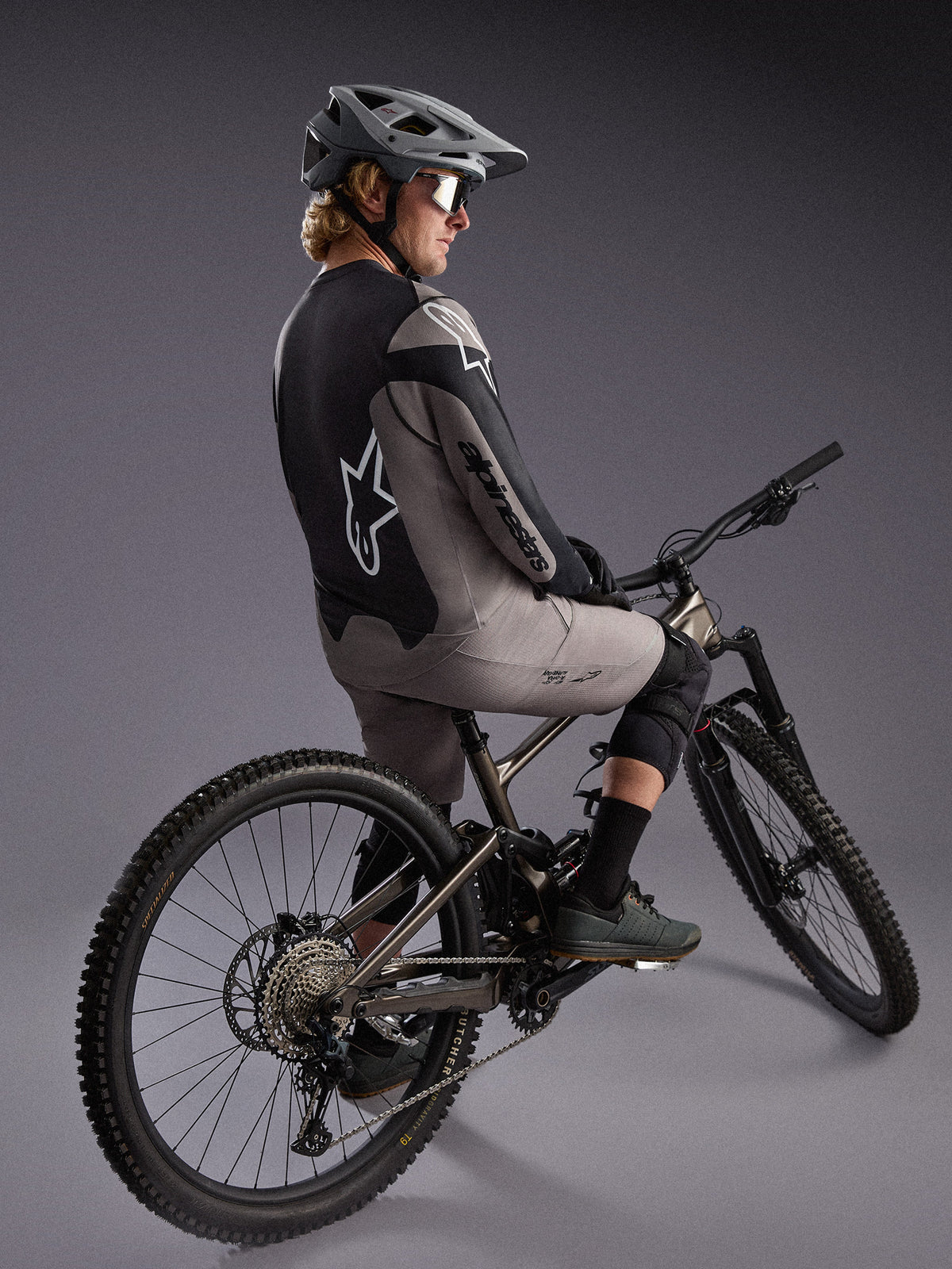 Camisola A-Dura Race cinzenta de manga comprida num ciclista de BTT, com logótipos Alpinestars e tecido elástico.