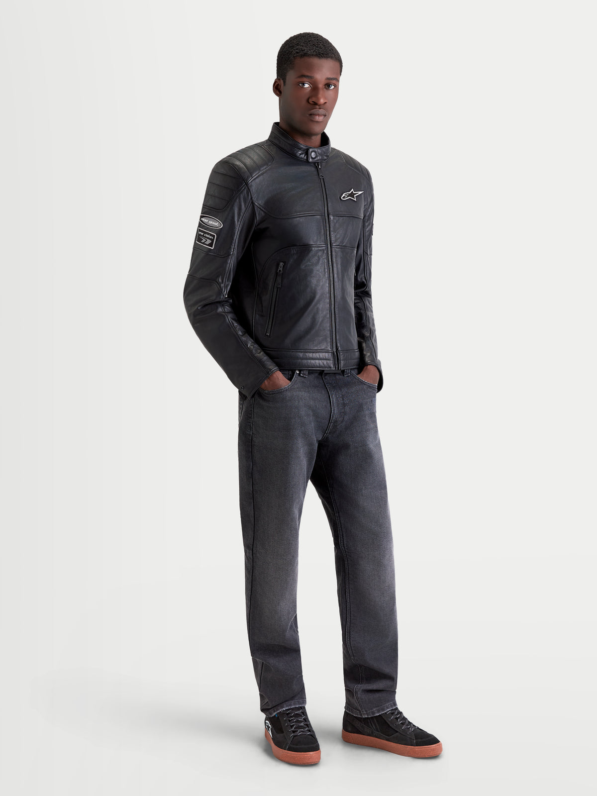 Alpinestars Revern casaco de couro, casaco de moto urbano em pele preto, vista frontal num modelo com calças de ganga escuras e sapatos de moto, com estilo heritage e construção em couro bovino de flor integral