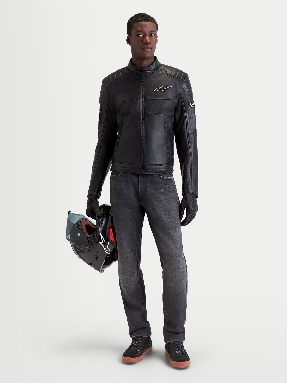 Alpinestars Revern, blusão de moto em couro para homem, preto, vista frontal num modelo a segurar um capacete e a vestir calças de ganga escuras, com construção em couro bovino envelhecido e estilo de herança biker