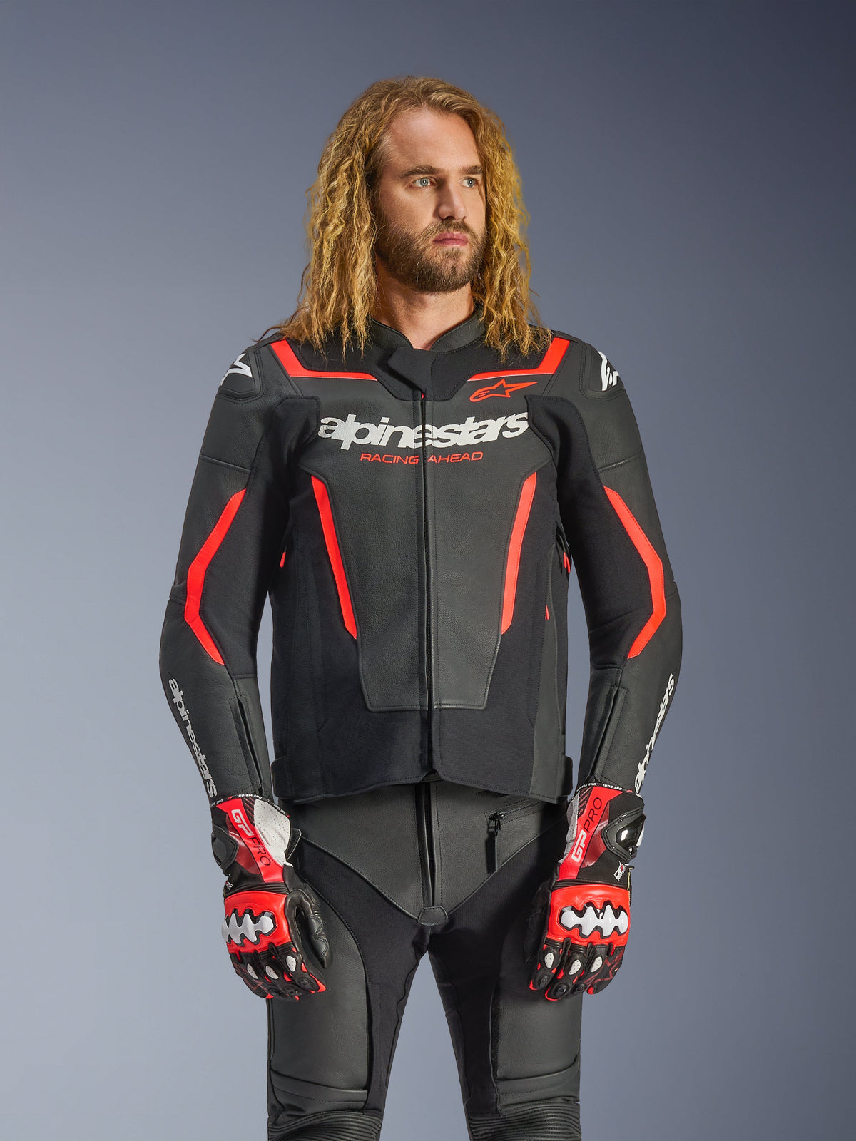 Alpinestars GP Force V2 Leather Jacket, Sport Motorcycle Jacket, Black and Red Fluo, worn by a male model with long wavy hair, featuring white Alpinestars logo and "RACING AHEAD" text, paired with matching leather pants and gloves, professional performance riding gear