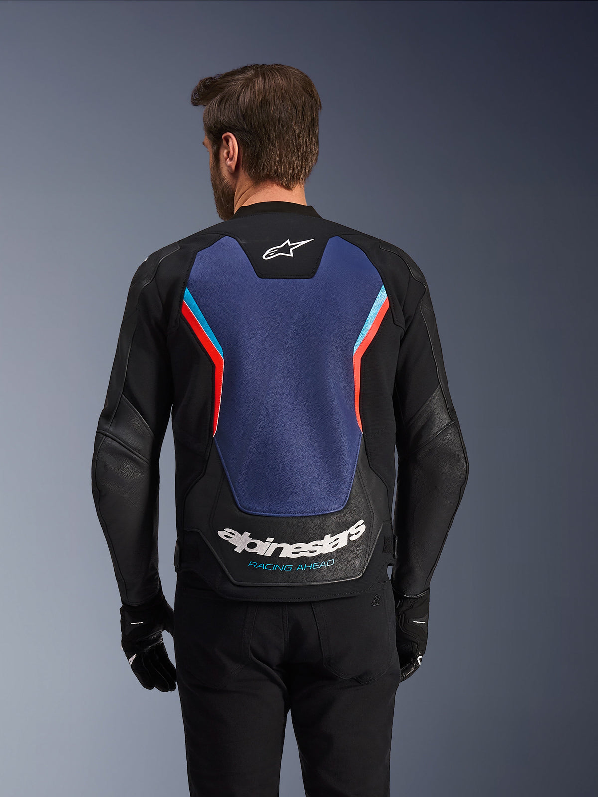 Vista traseira do Casaco de Pele Alpinestars GP Force V2 em Preto, Azul e Vermelho Fluo, com um grande painel azul nas costas, detalhes em vermelho fluorescente e azul claro, logótipo Alpinestars branco e marca 'Racing Ahead' na base, usado por um condutor com luvas e calças pretas.
