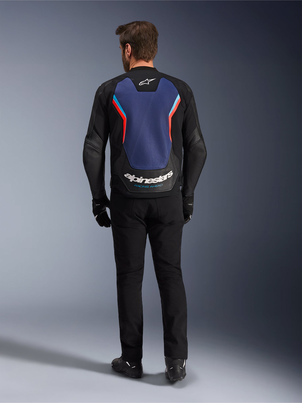 Casaco de Pele Alpinestars GP Force V2, casaco de motociclismo desportivo, preto, azul e vermelho fluo, vista traseira num modelo, com um grande painel traseiro azul com detalhes em vermelho e azul claro e a marca Alpinestars em branco na parte inferior das costas, combinado com calças e luvas de condução pretas.