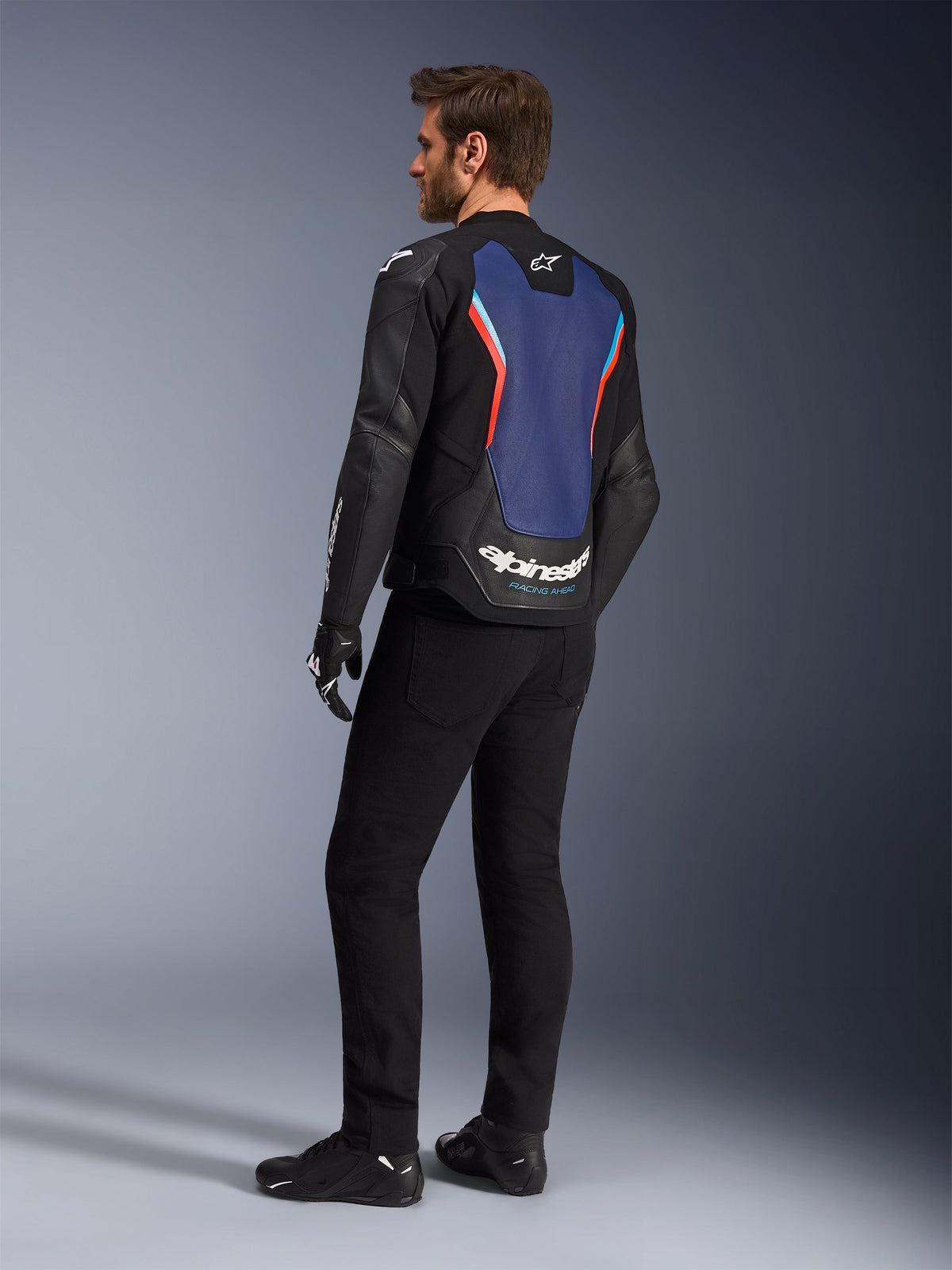 Vista traseira de três quartos de um modelo masculino a usar o Casaco de Pele Alpinestars GP Force V2 em Preto, Azul e Vermelho Fluorescente, destacando o painel traseiro azul com detalhes em vermelho fluorescente e logótipo branco, combinado com calças pretas e luvas de motociclista.