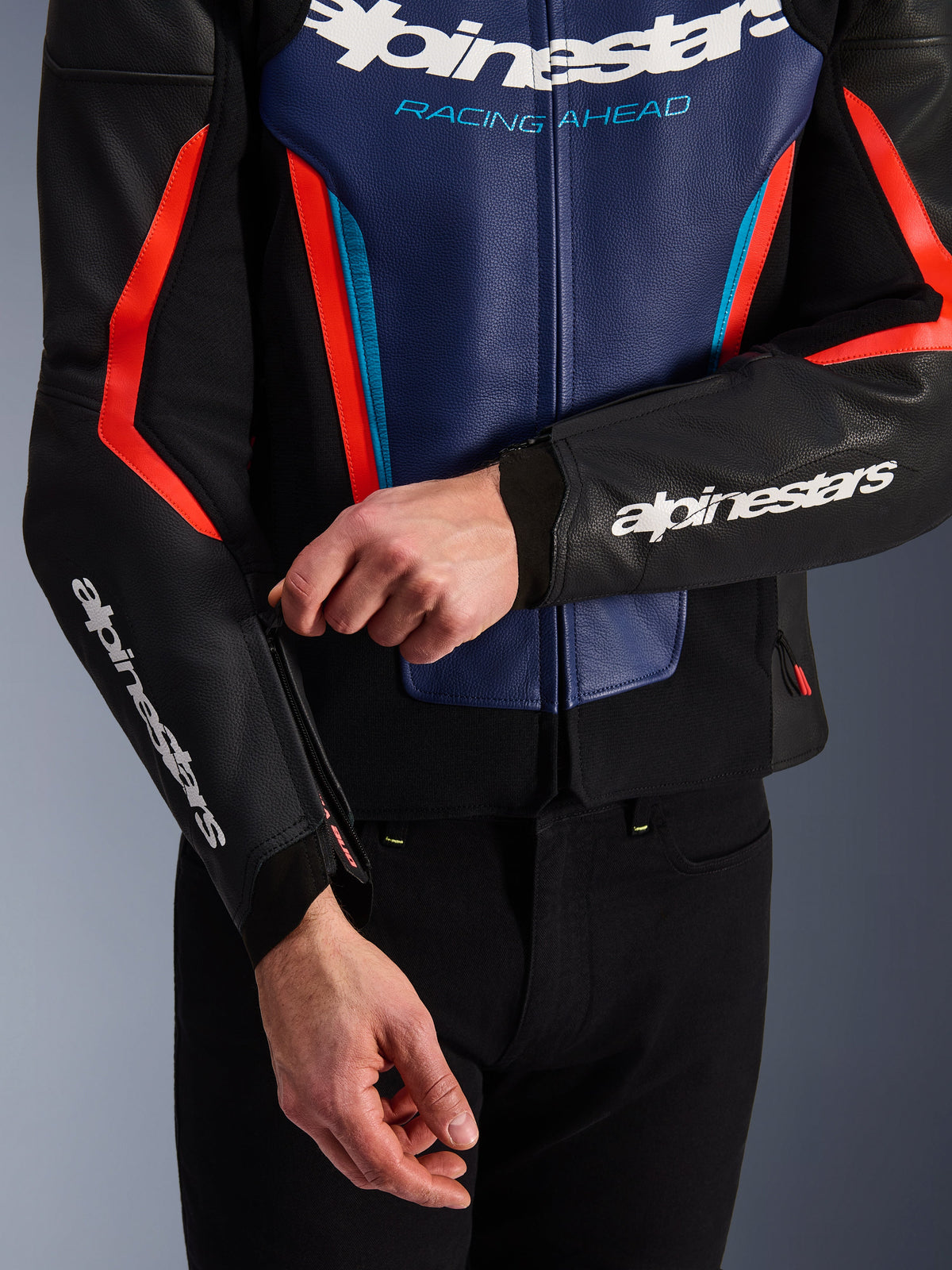 Casaco de Pele Alpinestars GP Force V2 em preto, azul e vermelho fluo, vista em pormenor de uma pessoa a ajustar o fecho do punho da manga, com logótipos Alpinestars brancos no antebraço e no peito, construção em pele bovina texturizada para condução de motos desportivas