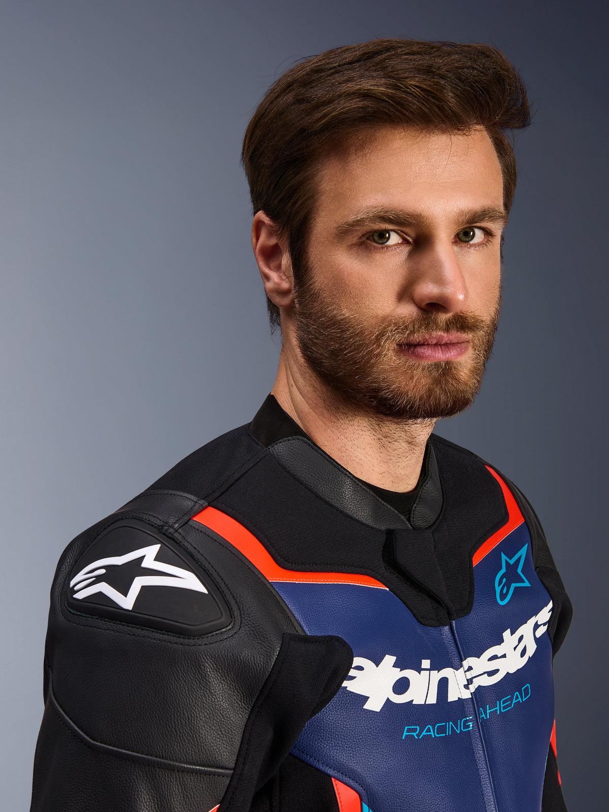Alpinestars GP Force V2 Leather Jacket, preto, azul e vermelho fluo, retrato em grande plano de um piloto masculino, com o logótipo Alpinestars branco nas proteções de ombro DFS pretas, painel de pele azul no peito com a marca branca e o texto 'Racing Ahead', detalhes em vermelho fluo, equipamento de motociclismo desportivo premium.