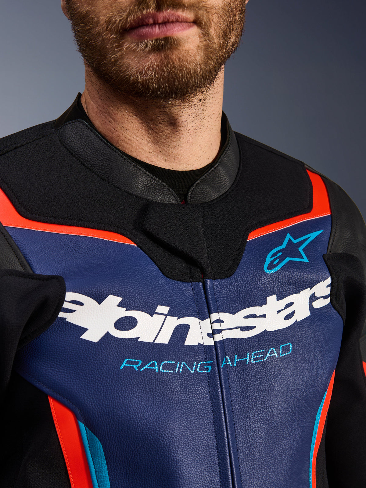 Casaco de Pele Alpinestars GP Force V2, vista detalhada do peito e gola, azul, preto e vermelho fluorescente, com o logótipo Alpinestars em branco e o texto 'RACING AHEAD' em azul claro, textura de pele premium com detalhes fluorescentes