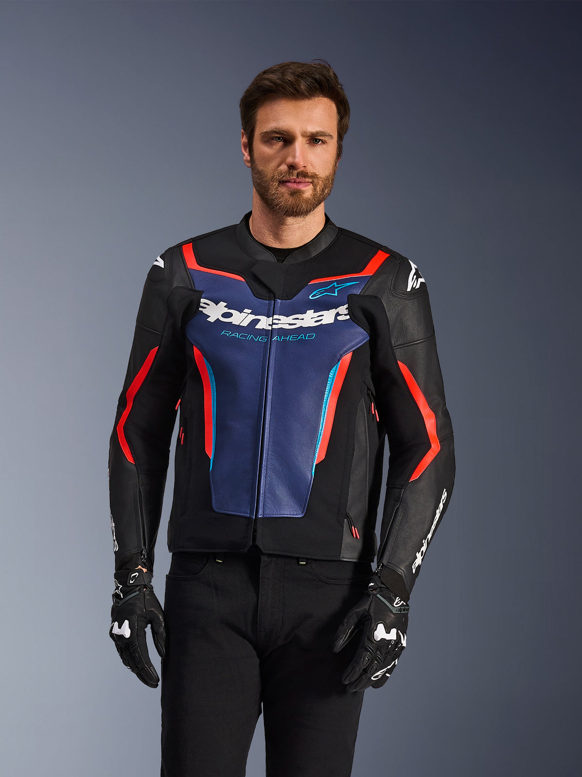 Casaco de Pele Alpinestars GP Force V2, preto, azul e vermelho fluo, vestido por um modelo masculino com luvas e calças pretas, com um painel azul no peito com o texto 'Alpinestars Racing Ahead', detalhes vermelhos e proteções externas nos ombros, equipamento de proteção para motociclismo desportivo