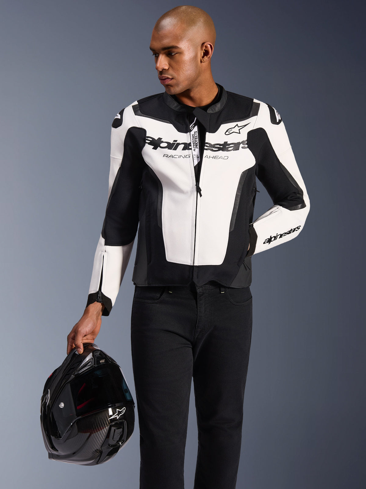 Model wearing the Alpinestars GP Force V2 Leather Jacket in White and Black, paired with black pants, holding a black motorcycle helmet, featuring a sporty multi-panel leather design with prominent Alpinestars branding on the chest and sleeves.