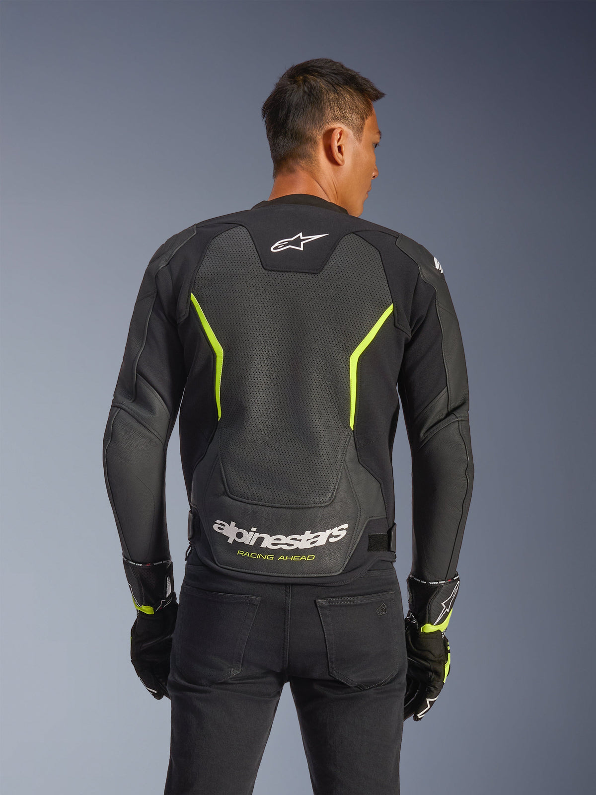 Casaco de Couro Alpinestars GP Force V2 Air, preto e amarelo néon, vista traseira de um piloto mostrando painéis extensos de couro perfurado para fluxo de ar, logótipo Alpinestars branco e texto "Racing Ahead" amarelo na zona lombar, equipamento desportivo de motociclismo para estrada