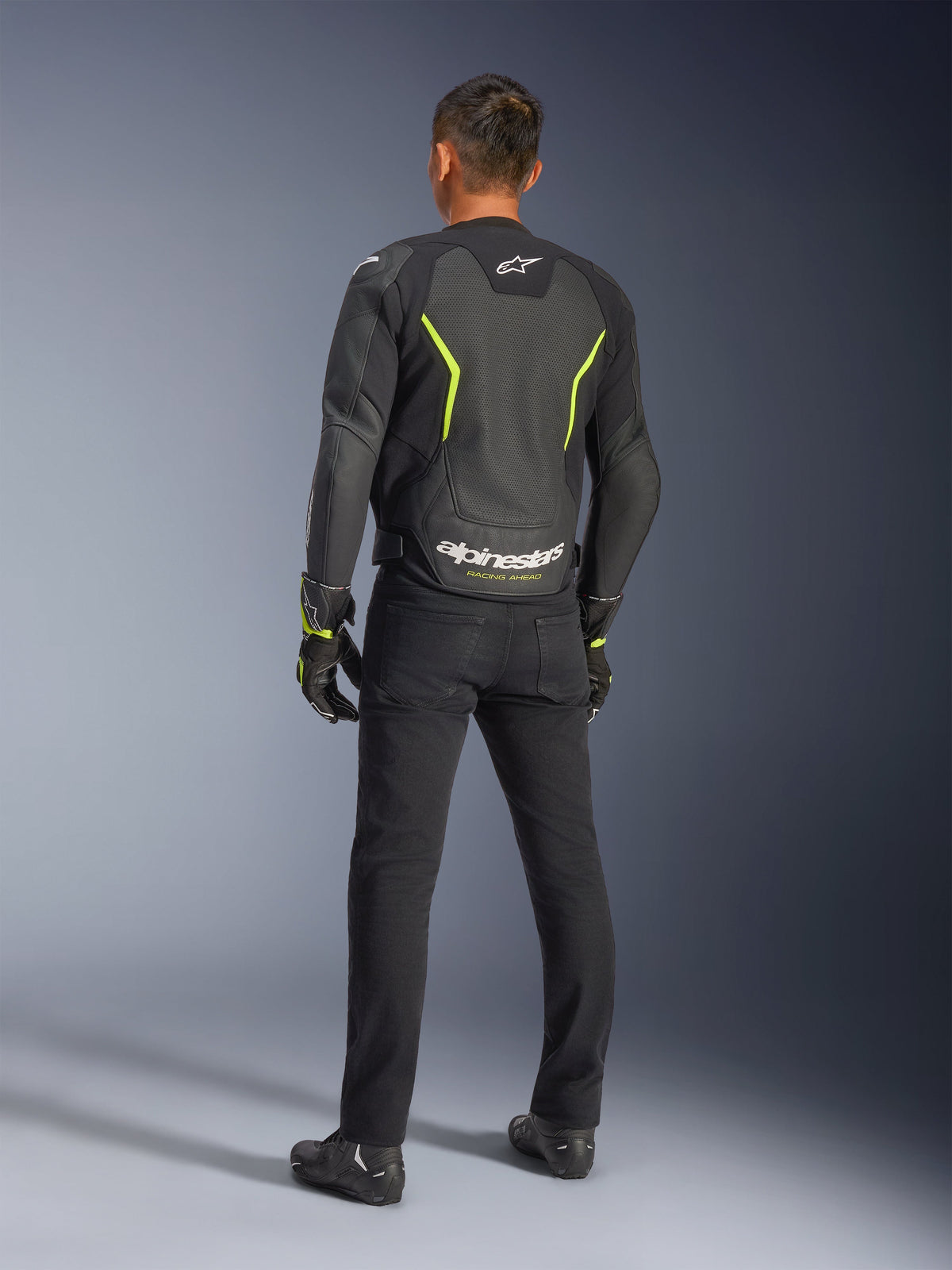 Casaco de Couro Alpinestars GP Force V2 Air, Preto e Amarelo Fluo, vista traseira de um condutor a usar o casaco de couro perfurado, com detalhes angulares em amarelo e logótipo Alpinestars branco em destaque com 'RACING AHEAD' na zona lombar, combinado com calças e botas de motociclismo pretas.