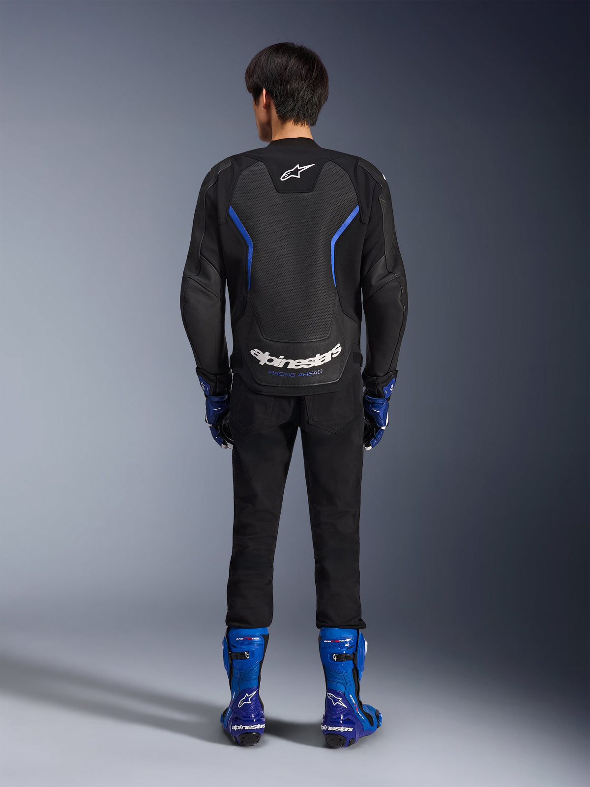 Casaco de Pele Alpinestars GP Force V2 Airflow, Preto e Azul Metálico, vista traseira a mostrar painéis de pele perfurada e logótipo branco, usado com botas de corrida azuis e calças pretas, equipamento de competição para motociclismo desportivo