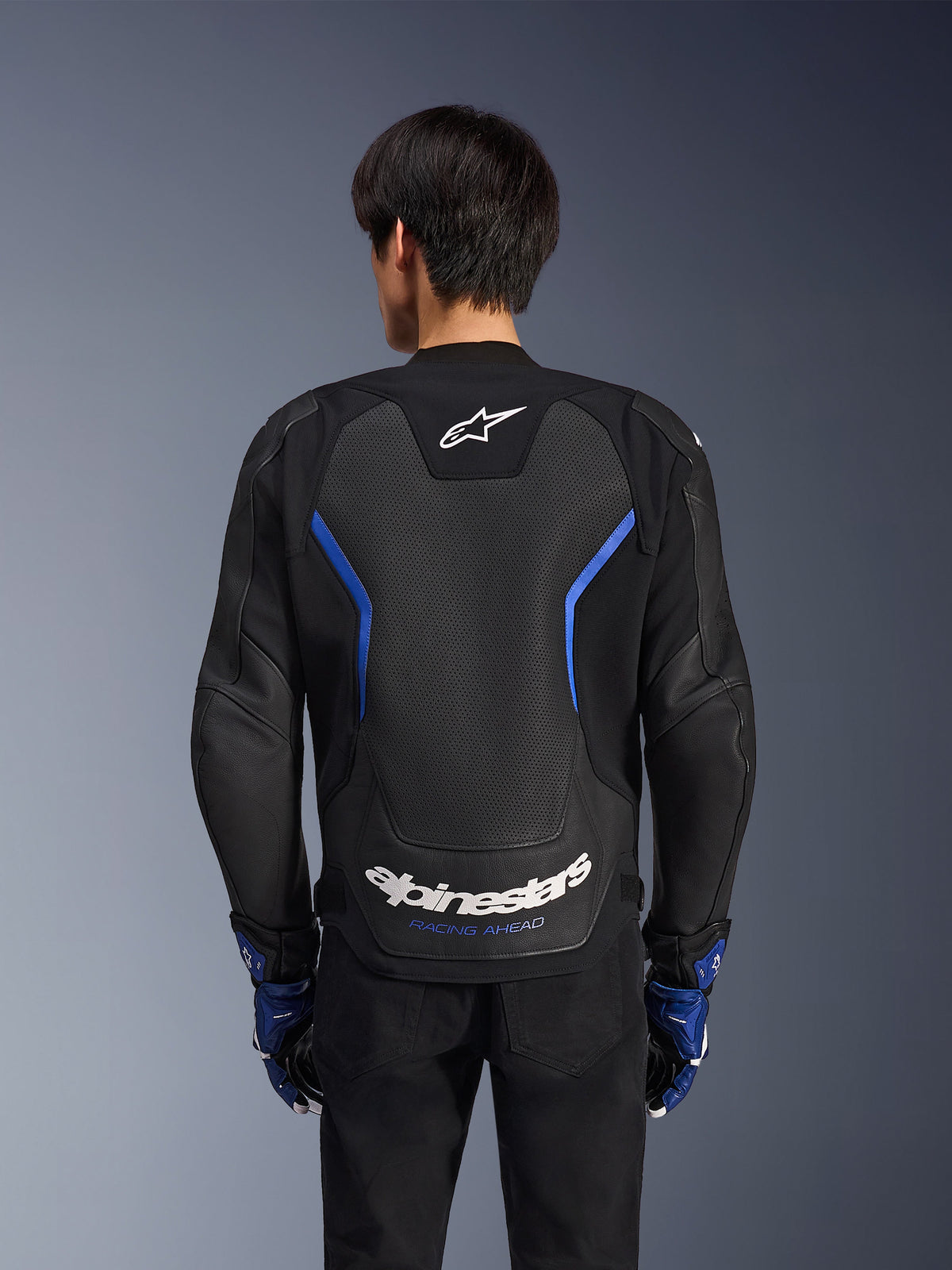 Alpinestars GP Force V2 Airflow Leather Jacket, preto e azul metálico, vista traseira, com um grande painel traseiro perfurado para ventilação, detalhes em azul e logótipo Alpinestars em branco, usado com luvas azuis a condizer e calças pretas para condução desportiva