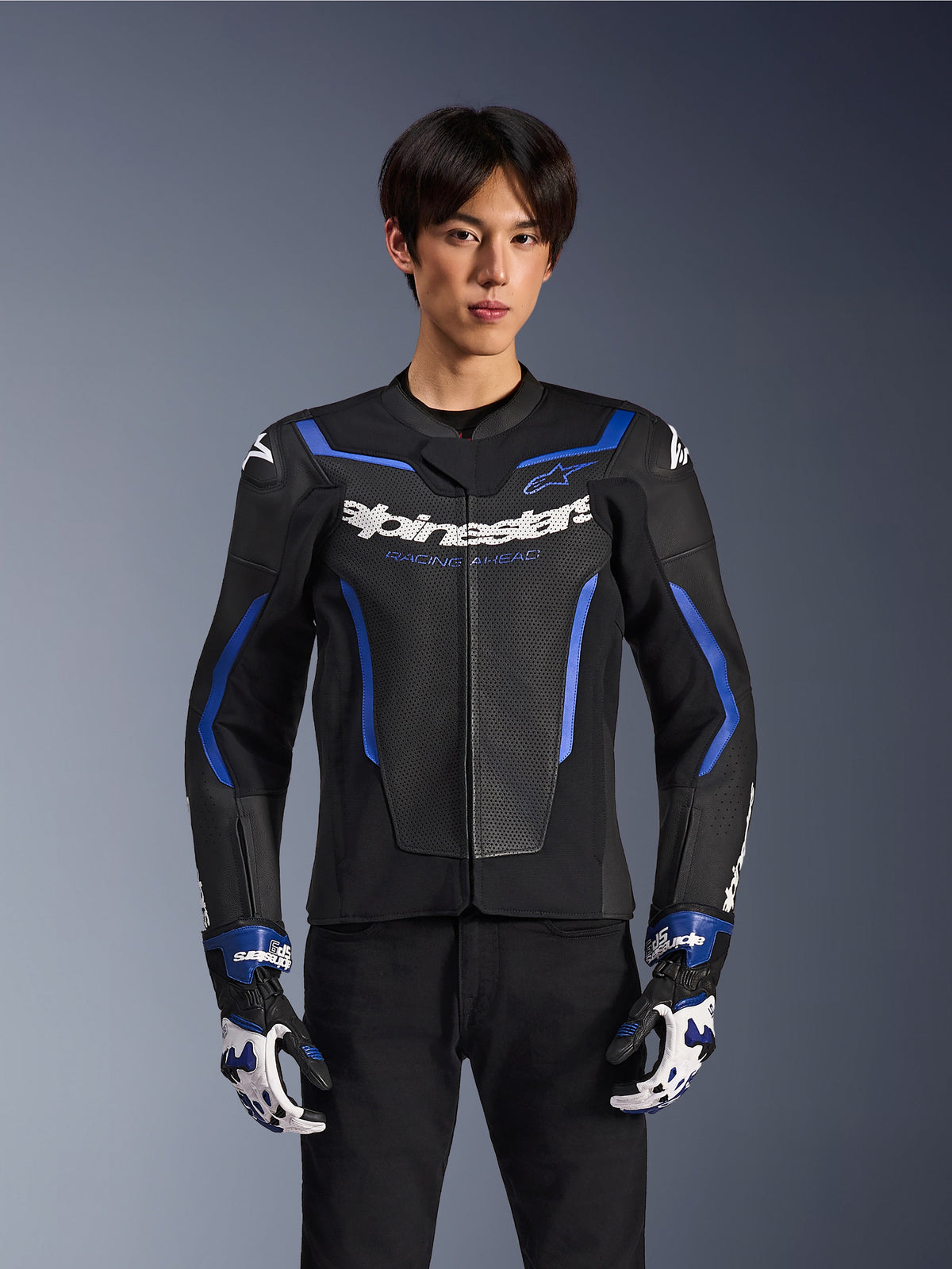 Casaco de Pele Alpinestars GP Force V2 Airflow, Black Metallic Blue, casaco de motociclista com pele perfurada e detalhes em azul, usado por um modelo com luvas de corrida a condizer e calças pretas, equipamento de condução desportiva profissional