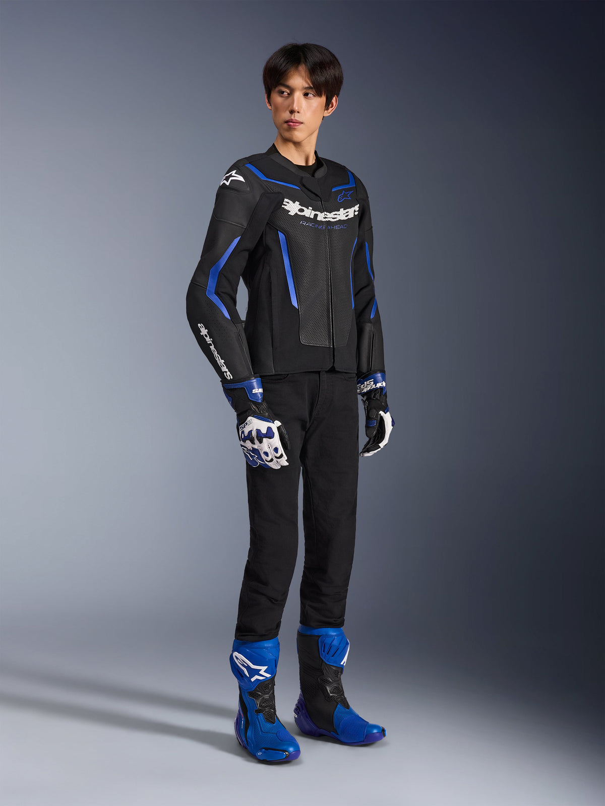 Condutor a usar o Casaco de Pele GP Force V2 Airflow em Preto e Azul Metálico, combinado com luvas de corrida a condizer e botas azuis, com pele perfurada e logótipos Alpinestars em destaque, equipamento profissional de motociclismo de estrada.