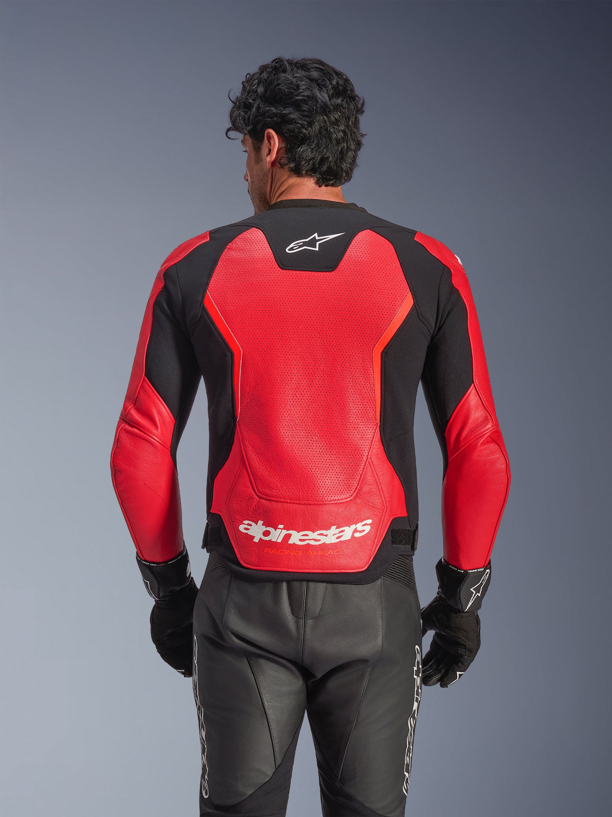 Alpinestars GP Force V2 Air Leather Jacket in Red and Black, rear view showing the perforated leather chassis for airflow, white Alpinestars branding on the lower back, and integrated stretch panels, worn with black leather motorcycle pants and gloves.