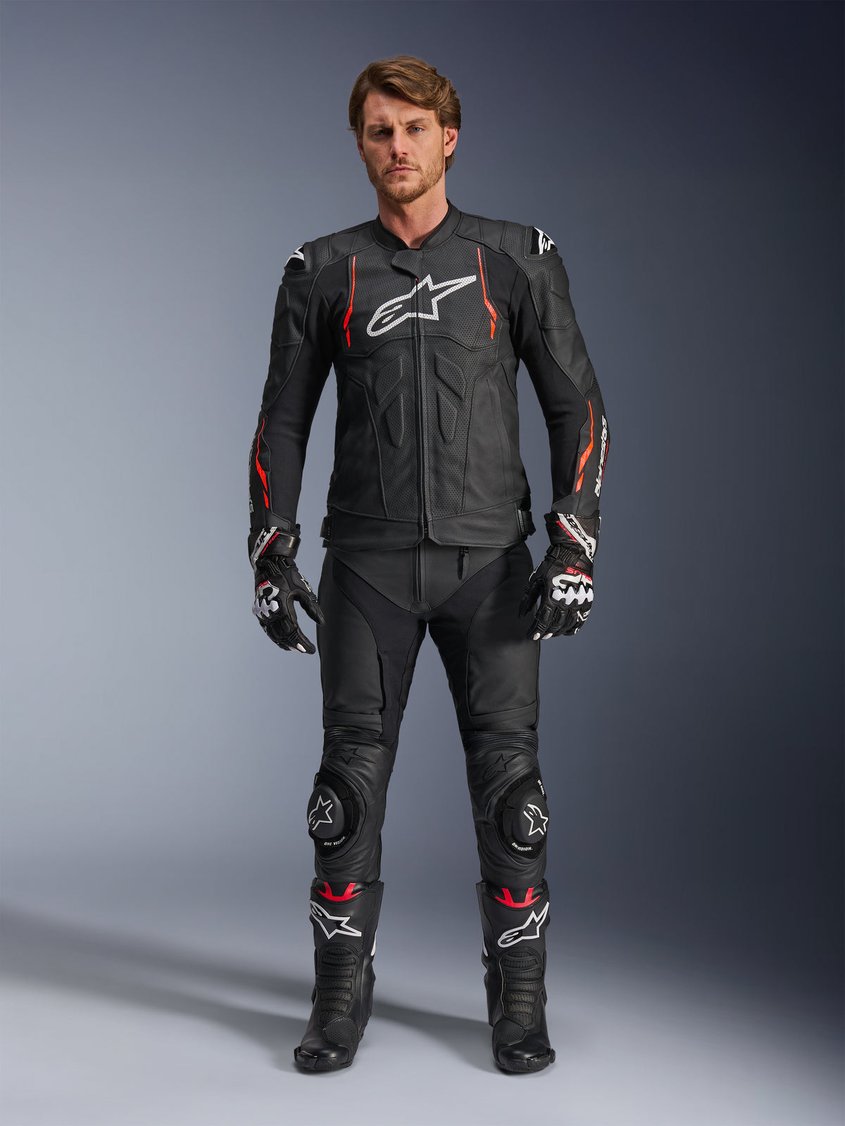 Motociclista masculino a usar o Casaco de Couro Alpinestars Dusk Airflow em Black Red Fluo, com painéis de couro perfurado e logótipo branco no peito, combinado com calças de couro pretas, sliders de joelho, luvas e botas de corrida, equipamento completo de motociclismo de estrada