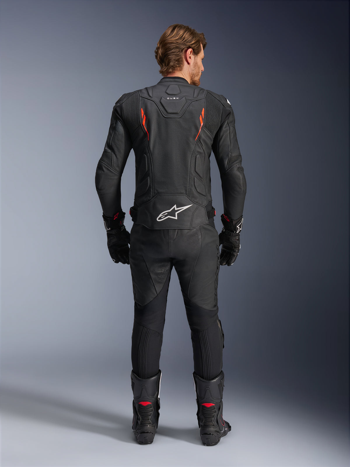 Alpinestars Dusk Airflow Leather Jacket, casaco de couro, preto e vermelho fluo, vista traseira com perfurações extensas e logótipo branco grande na parte inferior das costas, usado com equipamento de corrida em couro a condizer, incluindo calças, botas e luvas