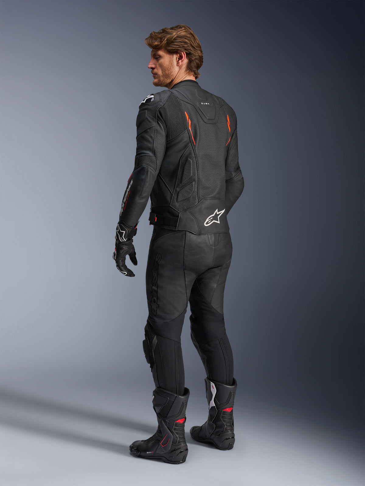 Casaco de Couro Alpinestars Dusk Airflow, preto com detalhes em Vermelho Fluo, vista traseira de um piloto com equipamento de motociclismo completo em couro, incluindo calças, luvas e botas de corrida pretas a condizer, com couro perfurado e logótipos Alpinestars brancos