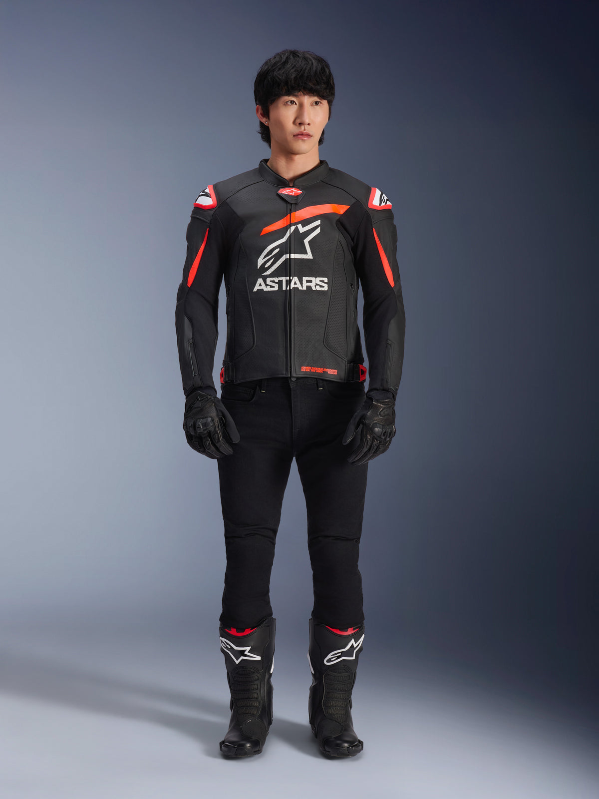 Modelo masculino a usar o Casaco de Couro Alpinestars GP Plus R V4 Airflow, preto com detalhes em vermelho fluo e branco, logótipo Astars branco de grandes dimensões no peito, combinado com calças técnicas, luvas e botas pretas, equipamento de motociclismo de alta performance num cenário de estúdio