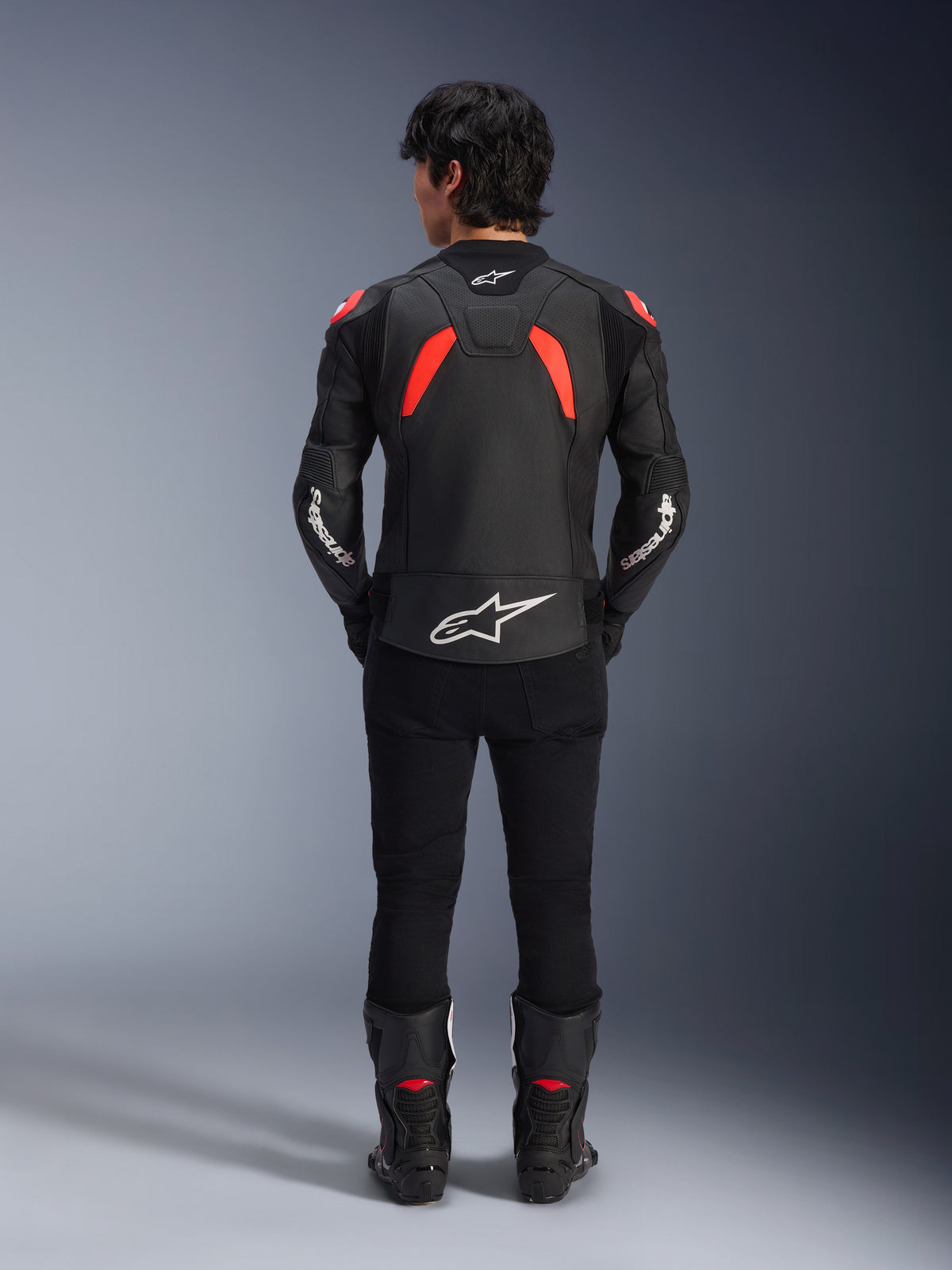 Jaqueta de Couro Alpinestars GP Plus R V4 Airflow, preto, vermelho fluo e branco, vista traseira num modelo, com painéis de couro perfurados e logótipos brancos destacados, combinada com calças e botas de motociclismo pretas