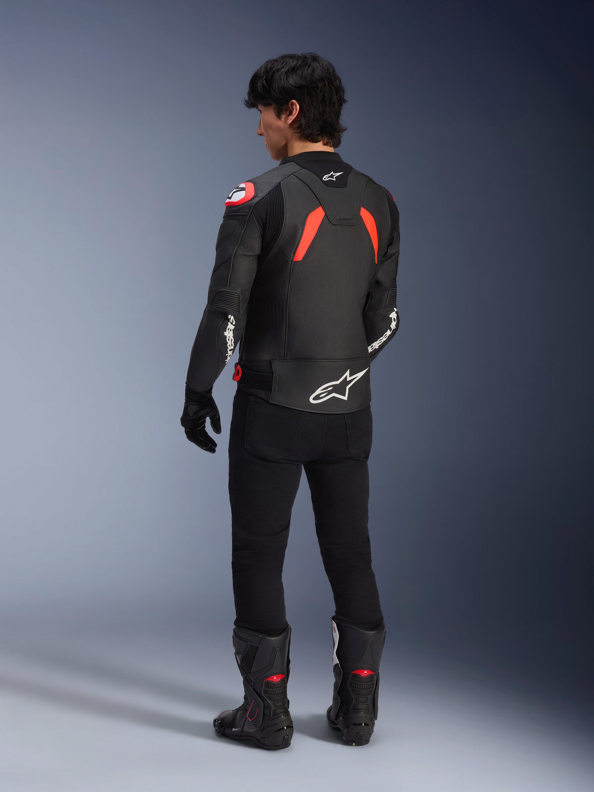 Vista traseira de um modelo a usar a Jaqueta de Couro Alpinestars GP Plus R V4 Airflow, preta com detalhes em Vermelho Fluo e Branco, usada com calças pretas e botas de motociclismo pretas, apresentando logótipos brancos proeminentes nas costas e mangas, construção em couro de estilo desportivo