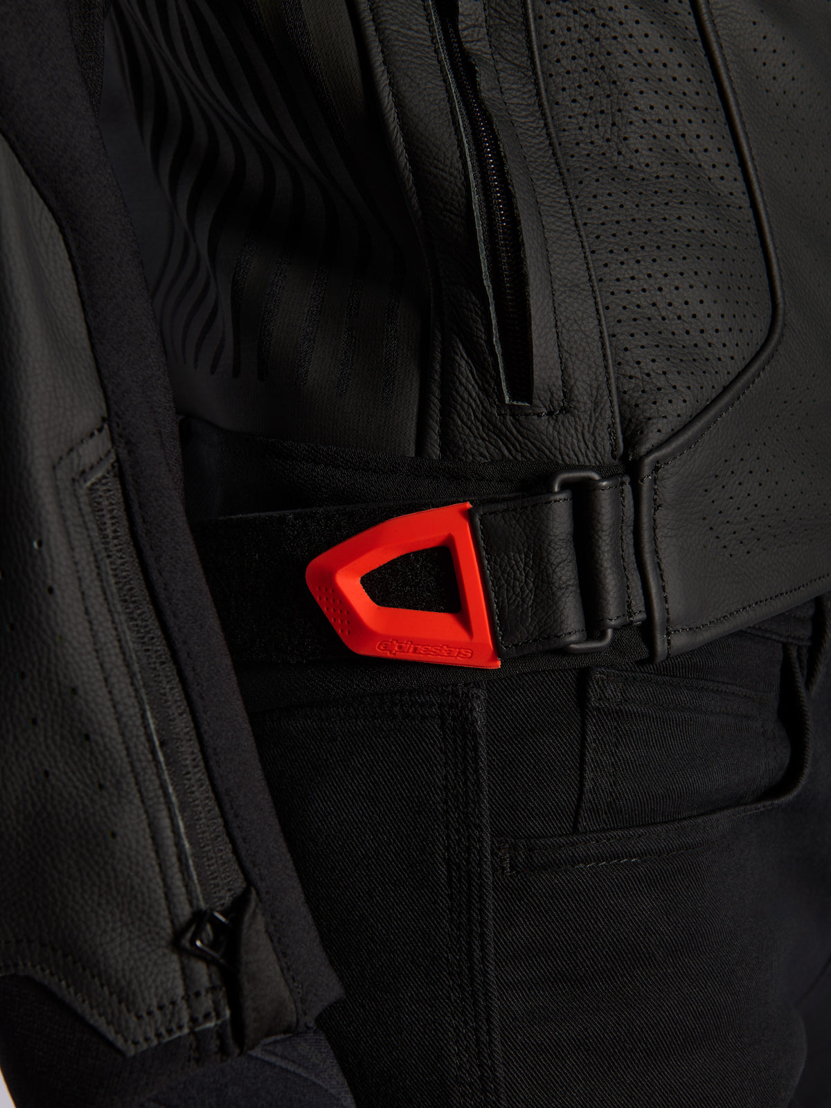 Alpinestars GP Plus R V4 Airflow Leather Jacket, pormenor de casaco de couro preto perfurado com correia de ajuste de cintura em gancho e laço vermelho fluo vibrante, bolso lateral com fecho e painéis texturizados ergonómicos.