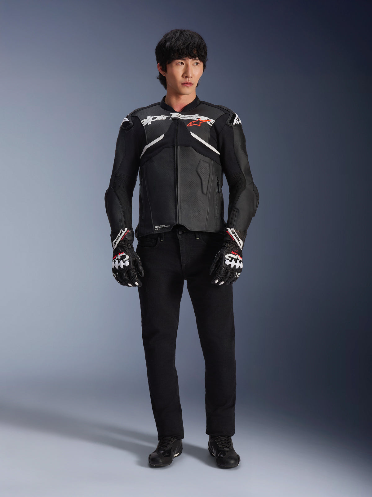 Modelo a usar o Casaco de Couro Alpinestars Atem V5 em preto e branco, com a marca Alpinestars em branco e o logótipo da estrela em vermelho no peito, combinado com calças pretas e luvas de corrida pretas e brancas, equipamento de motociclismo de alta performance