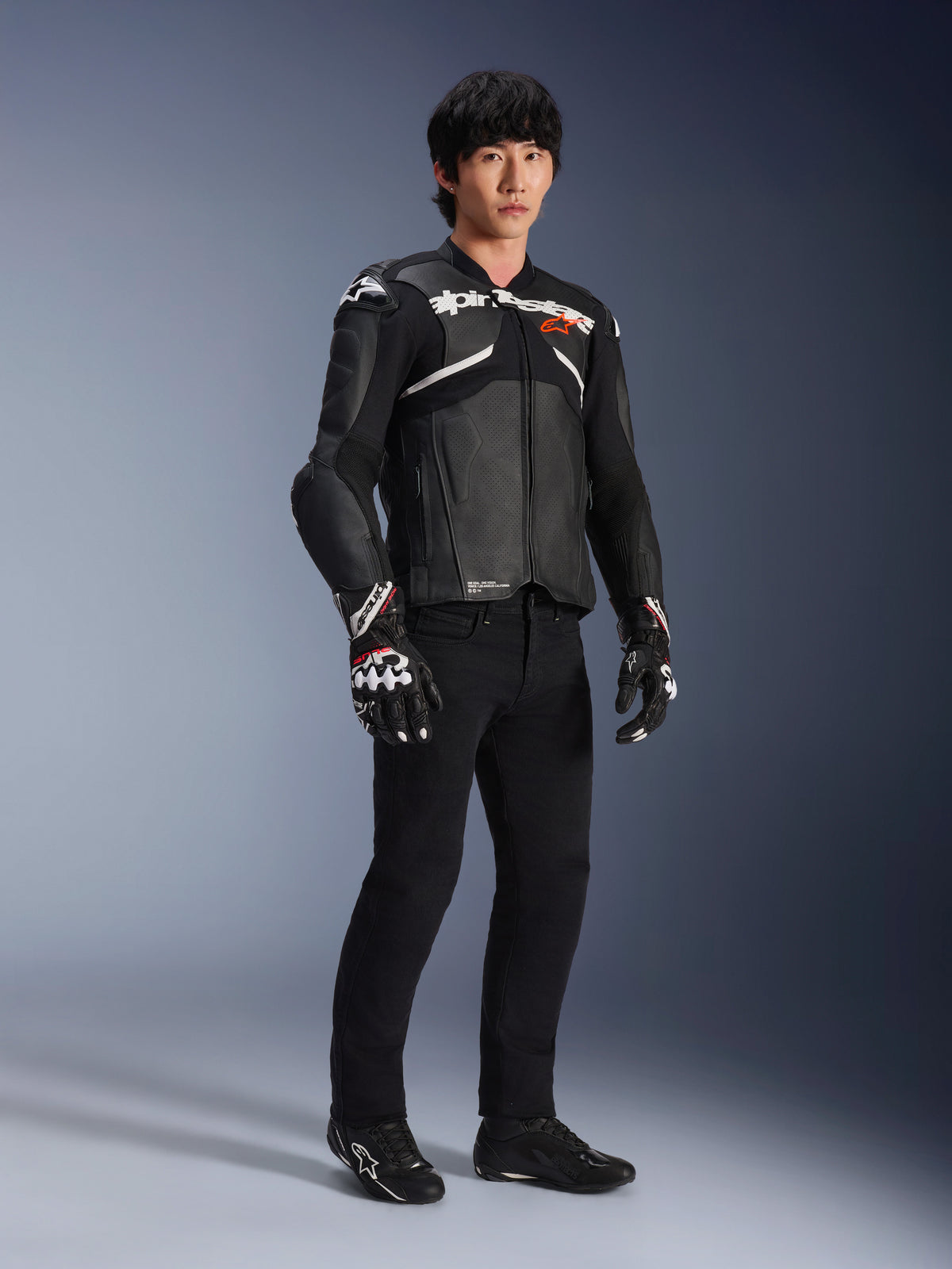 Jaqueta de Couro Alpinestars Atem V5, casaco de corrida para motociclismo, preto e branco, usado com calças pretas e luvas a condizer, com a marca Alpinestars em branco no peito e proteção nos ombros.