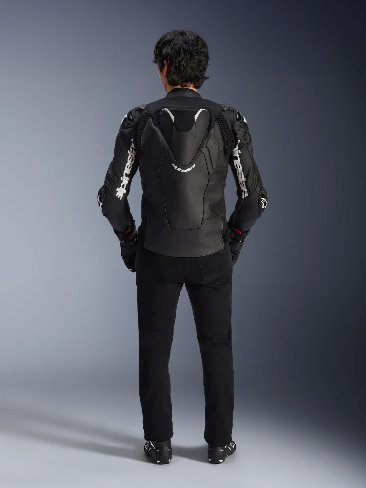 Jaqueta de Couro Alpinestars Atem V5, jaqueta de corrida para motociclismo, preto e branco, vista traseira no piloto, com corcova aerodinâmica e logótipos Alpinestars brancos nas mangas e nas costas, construção técnica em couro, usado com calças pretas e botas de motociclismo