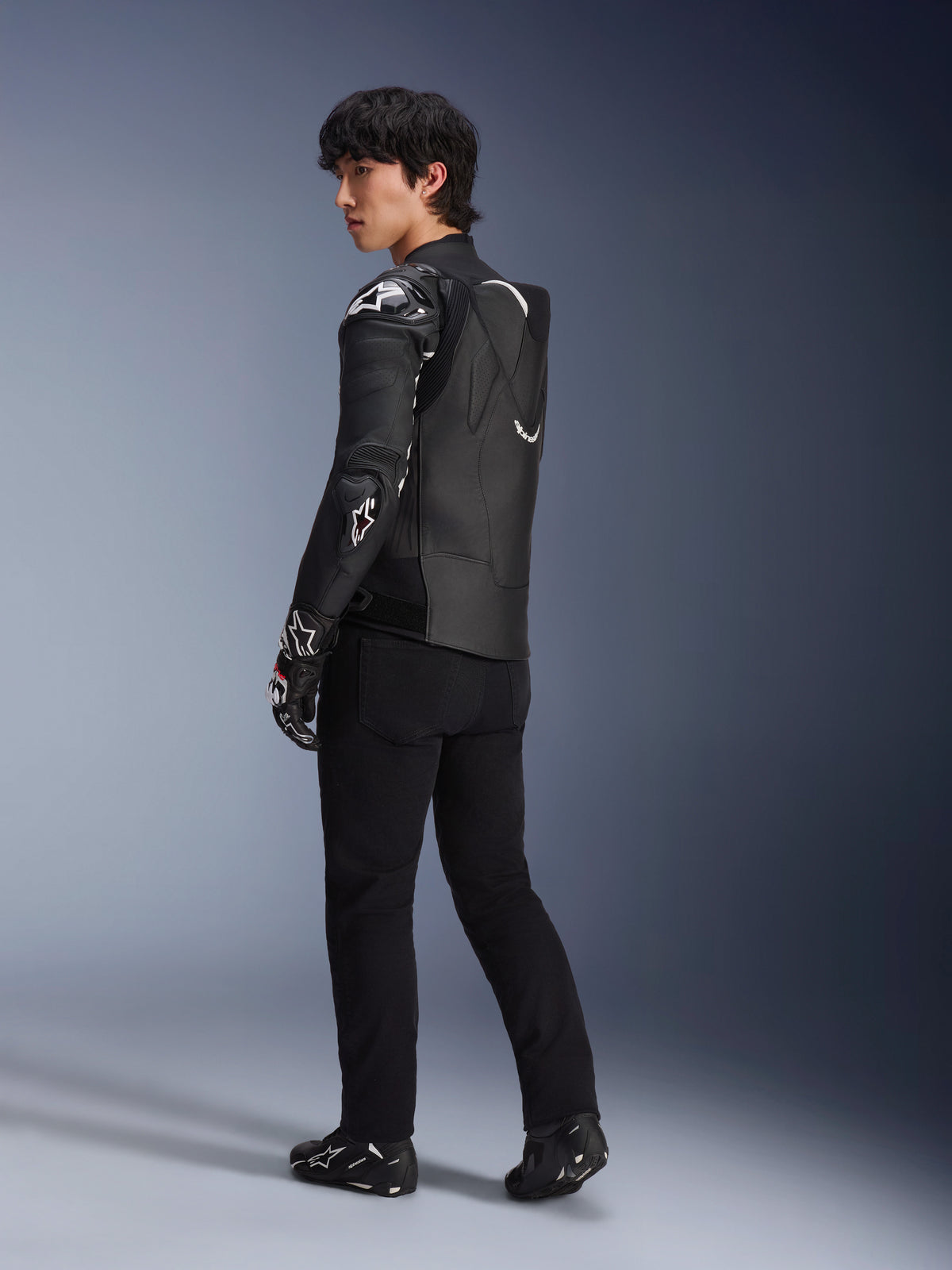 Modelo masculino a usar o Casaco de Couro Alpinestars Atem V5 em preto e branco, vista traseira de três quartos mostrando a corcova aerodinâmica e os protetores DFS RR, combinado com calças pretas, luvas de corrida e botas, equipamento profissional de corrida de motociclismo