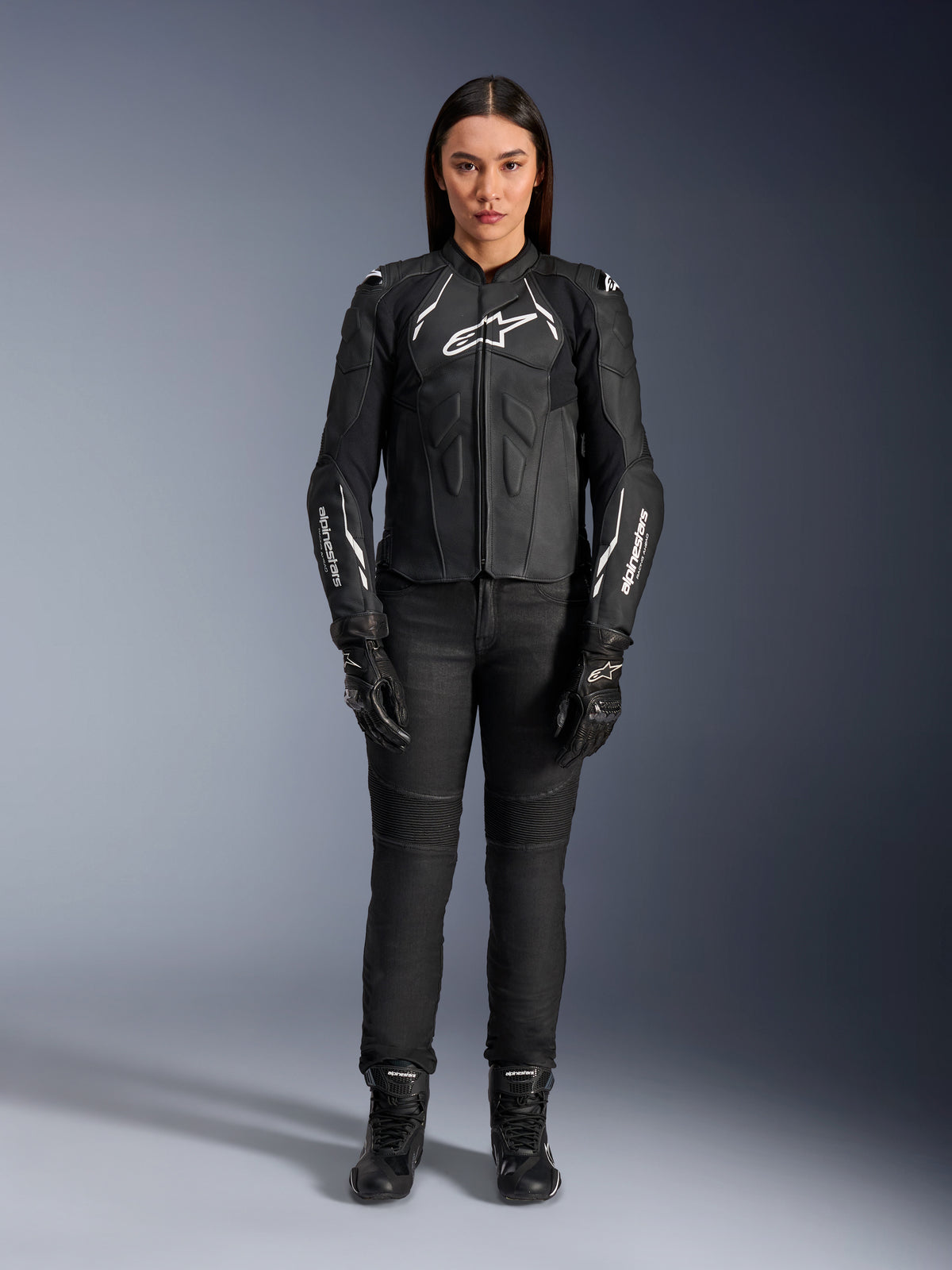 Casaco de Couro Feminino Alpinestars Stella Dusk, preto e branco, usado por uma modelo com calças e botas pretas a condizer, vista frontal, com logótipos Alpinestars brancos no peito e nas mangas, equipamento de proteção em couro bovino premium para mulher