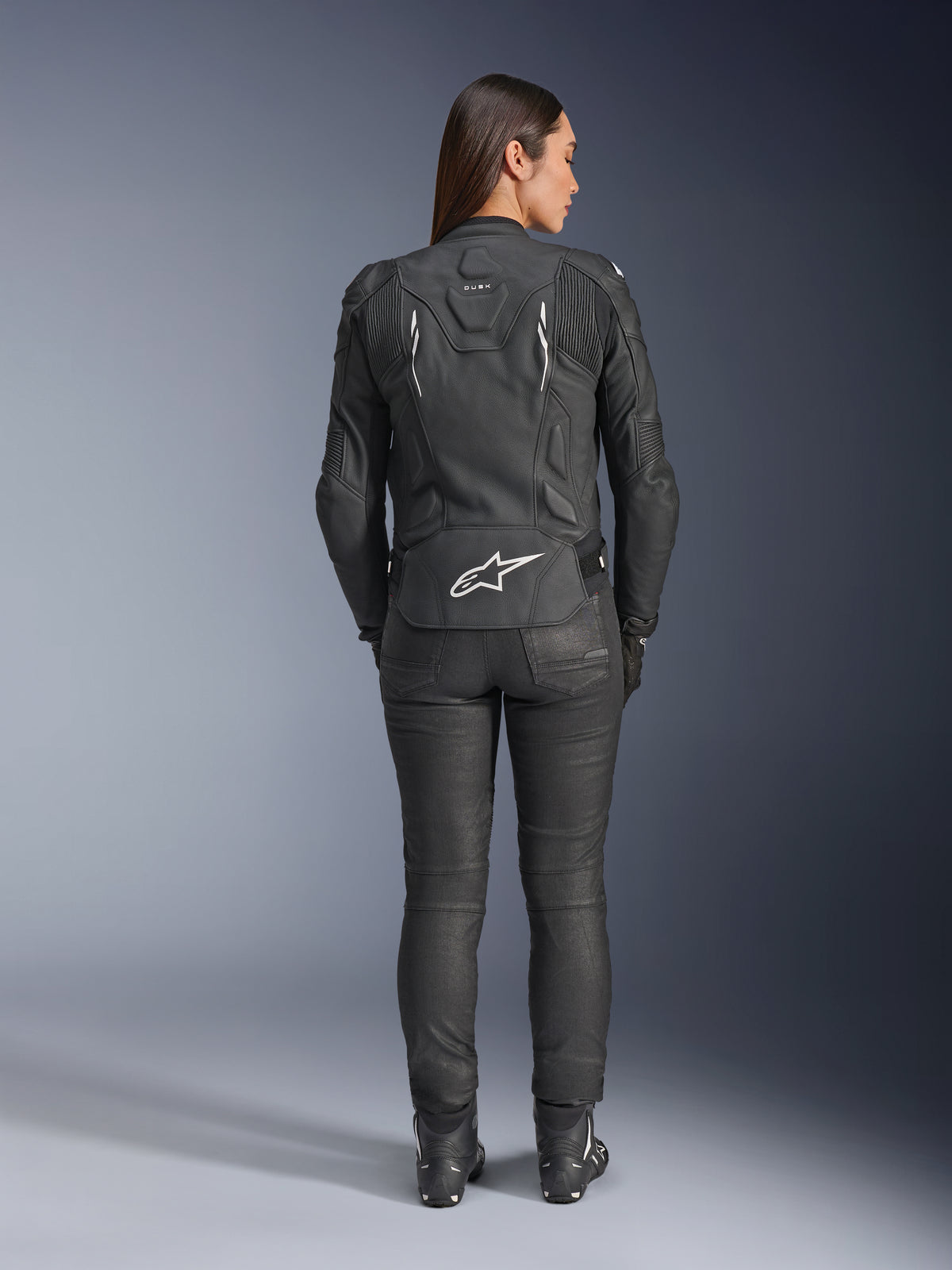 Casaco de Couro Feminino Alpinestars Stella Dusk, preto, vista traseira com o logótipo Alpinestars branco na parte inferior das costas e marca 'DUSK' na parte superior, usado com calças e botas de motociclismo pretas, realçando o ajuste feminino e a construção em couro protetor.