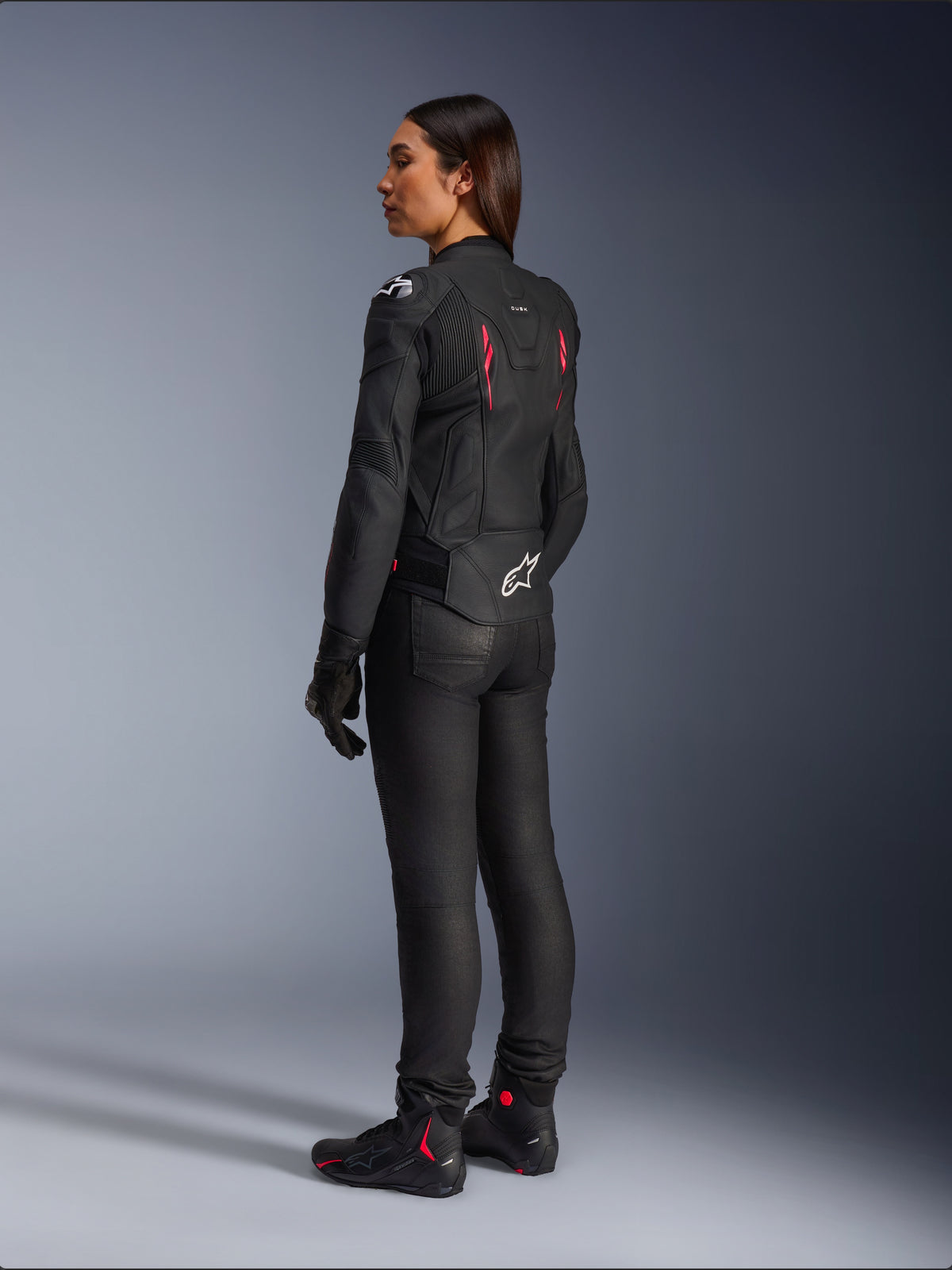 Woman Stella Dusk Leather Jacket, Black Diva Pink White, rear three-quarter view on a rider, worn with black riding pants and black boots, featuring sleek leather construction with pink accents and a white Alpinestars logo on the lower back.