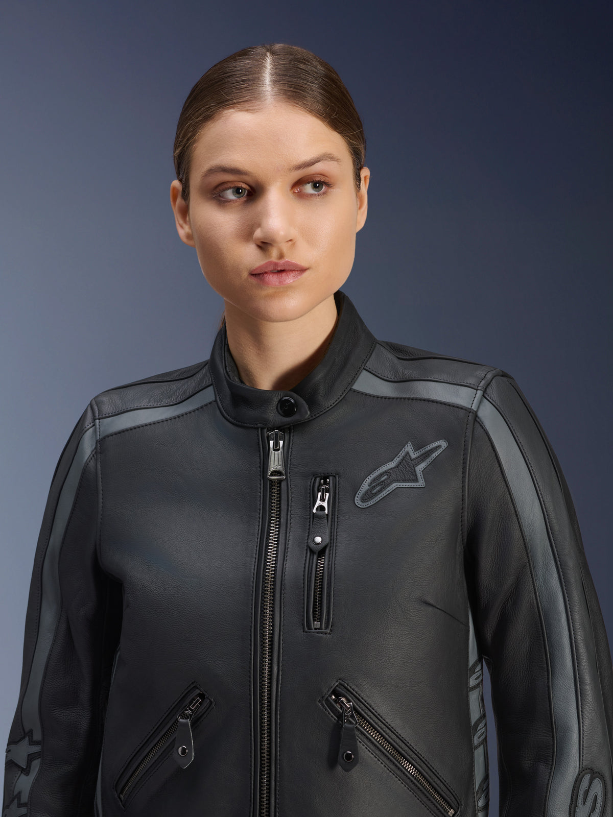 Modelo feminina a usar a Jaqueta de Couro Stella Dyno da Alpinestars em Black Tar Gray Dark Gray, jaqueta de motociclismo em couro bovino premium com design heritage, apresentando detalhes cinzentos nos ombros e vários bolsos com fecho de correr, com um corte especificamente feminino.