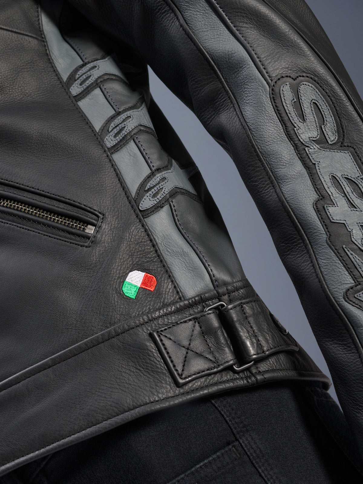 Detalhe da Jaqueta de Couro Alpinestars Stella Dyno, couro bovino preto e cinzento Tar, grande plano com pequeno bordado da bandeira italiana, fivela de cintura ajustável, painéis de couro cinzento com logótipos Alpinestars em relevo e marca Stella na manga