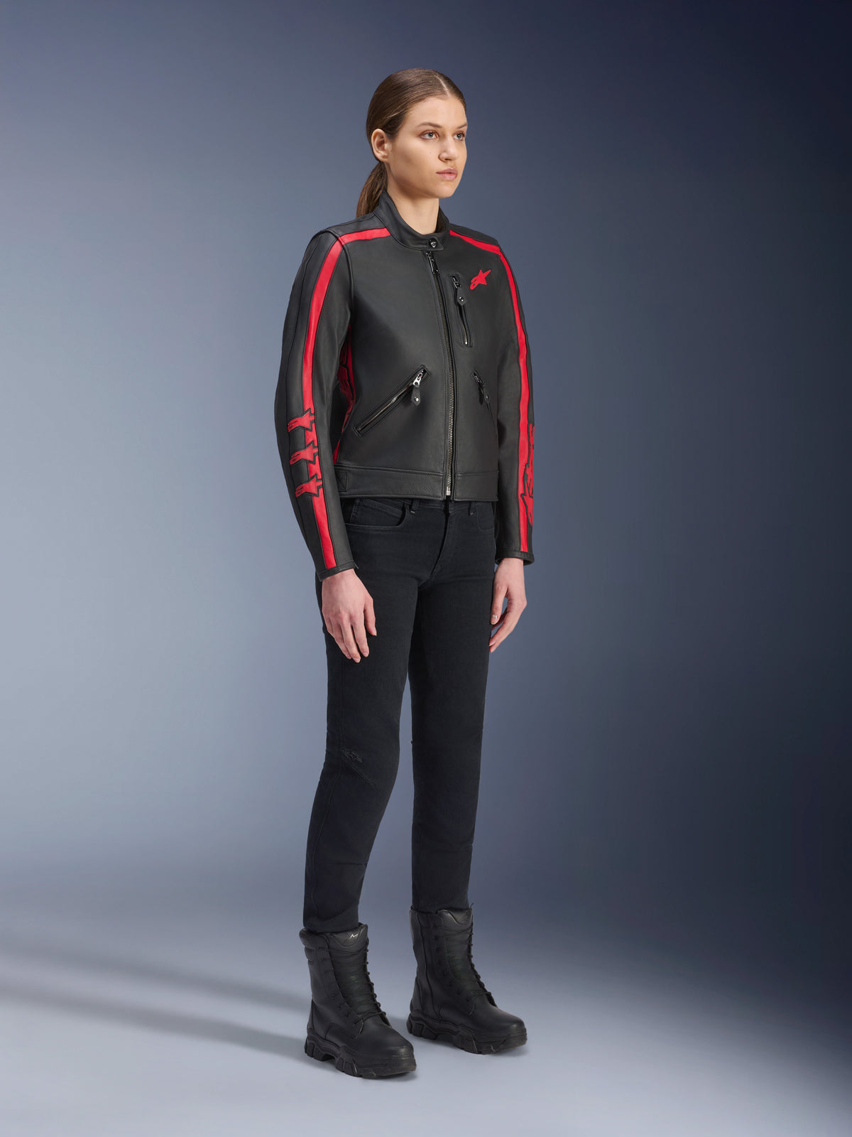 Modelo feminina a usar o casaco de pele Alpinestars Stella Dyno em Black Haute Red, combinado com calças e botas pretas, casaco de motociclismo em pele de estilo clássico com riscas vermelhas nos braços e logótipo vermelho, equipamento de condução urbana e de estrada para mulher