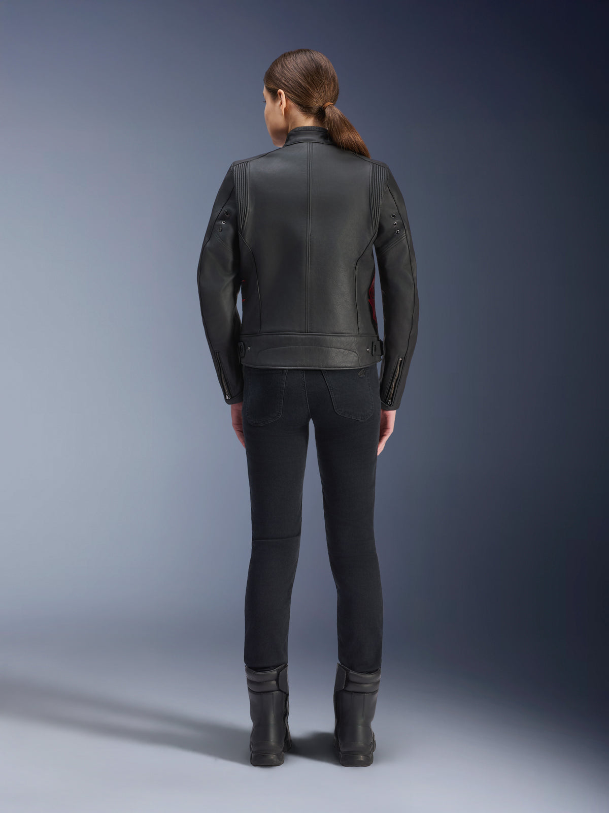 Vista traseira de uma mulher a usar o casaco de couro Alpinestars Stella Dyno, preto com pormenores em vermelho Haute Red, combinado com calças pretas justas e botas pretas, design de casaco de motociclismo em couro de estilo heritage moderno.