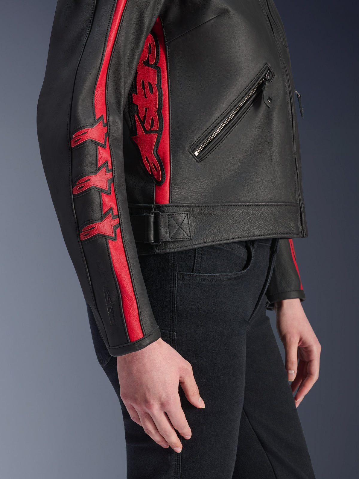 Casaco de Couro Alpinestars Stella Dyno, casaco de motociclismo para mulher, preto e vermelho Haute Red, vista lateral numa modelo, com riscas de corrida vermelhas e logótipos de estrela na manga, grande logótipo vermelho na lateral, couro bovino premium, estilo de condução urbana