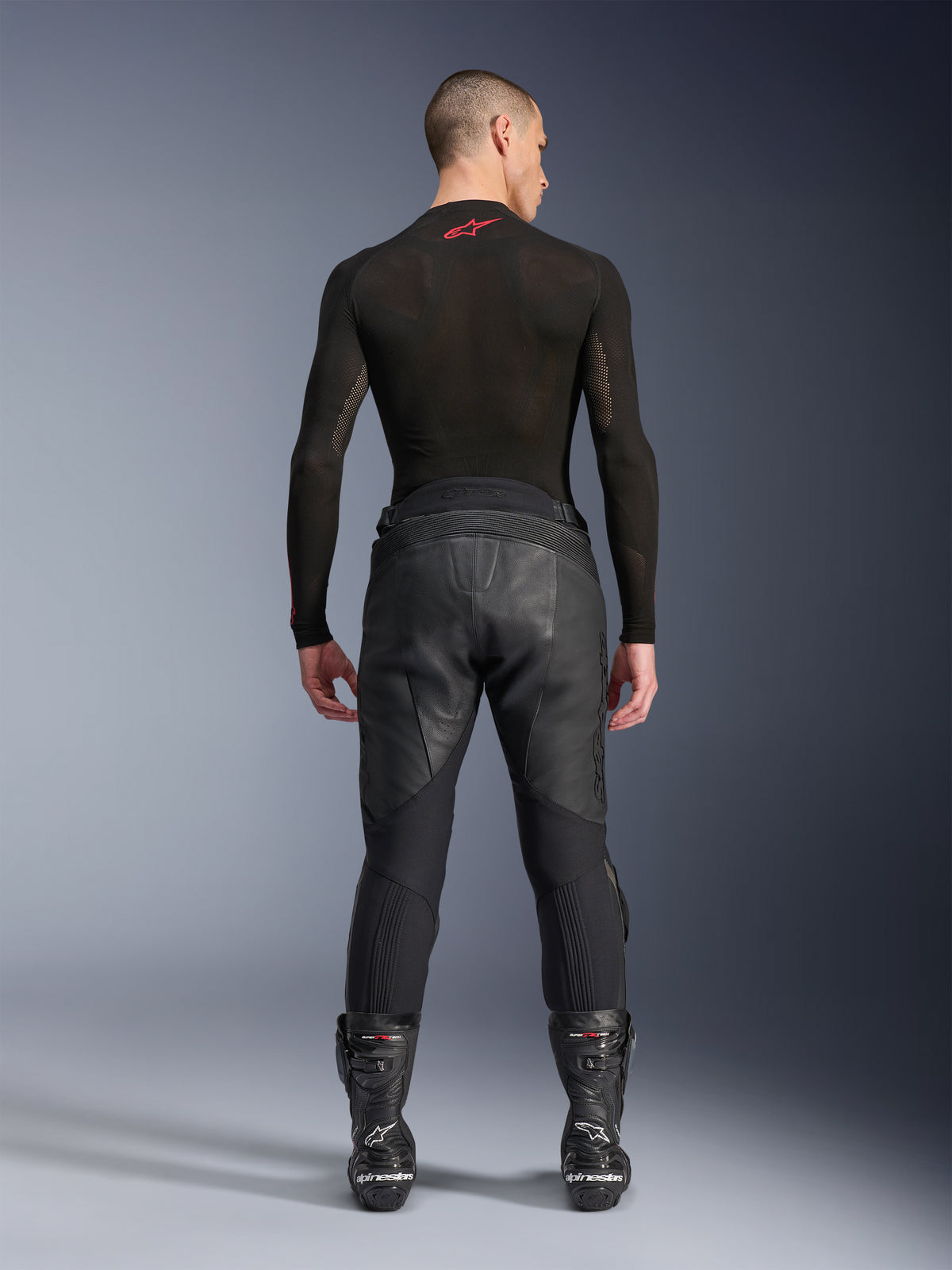 Alpinestars Missile V3 Leather Pants, Black, rear view on model wearing a black long-sleeve compression shirt and racing boots, premium leather construction with high-waist overlap and stretch zones, professional motorcycle racing gear