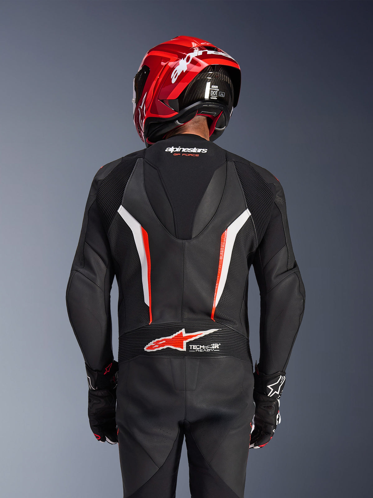 Alpinestars GP Force V2 Leather Suit 1-Piece, Black White and Red Fluo, back view of rider wearing red racing helmet and gloves, featuring perforated leather panels, aerodynamic speed hump with Alpinestars branding, and Tech-Air Ready logo on the lower back, professional motorcycle racing gear.