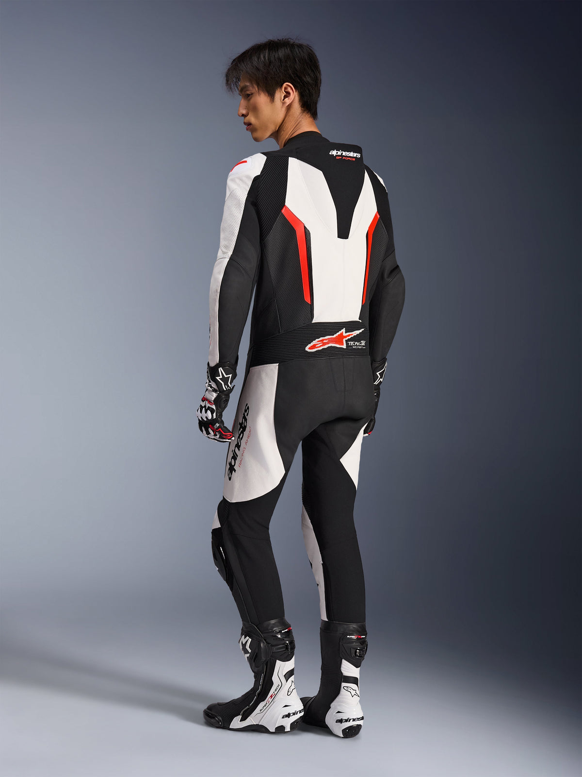 Motociclista a usar o fato de pele Alpinestars GP Force V2 - 1 peça em Branco, Preto e Vermelho Fluo, vista lateral traseira que mostra a corcova aerodinâmica e os painéis de pele perfurada, combinado com botas e luvas de corrida a condizer, concebido para pista e condução de performance.