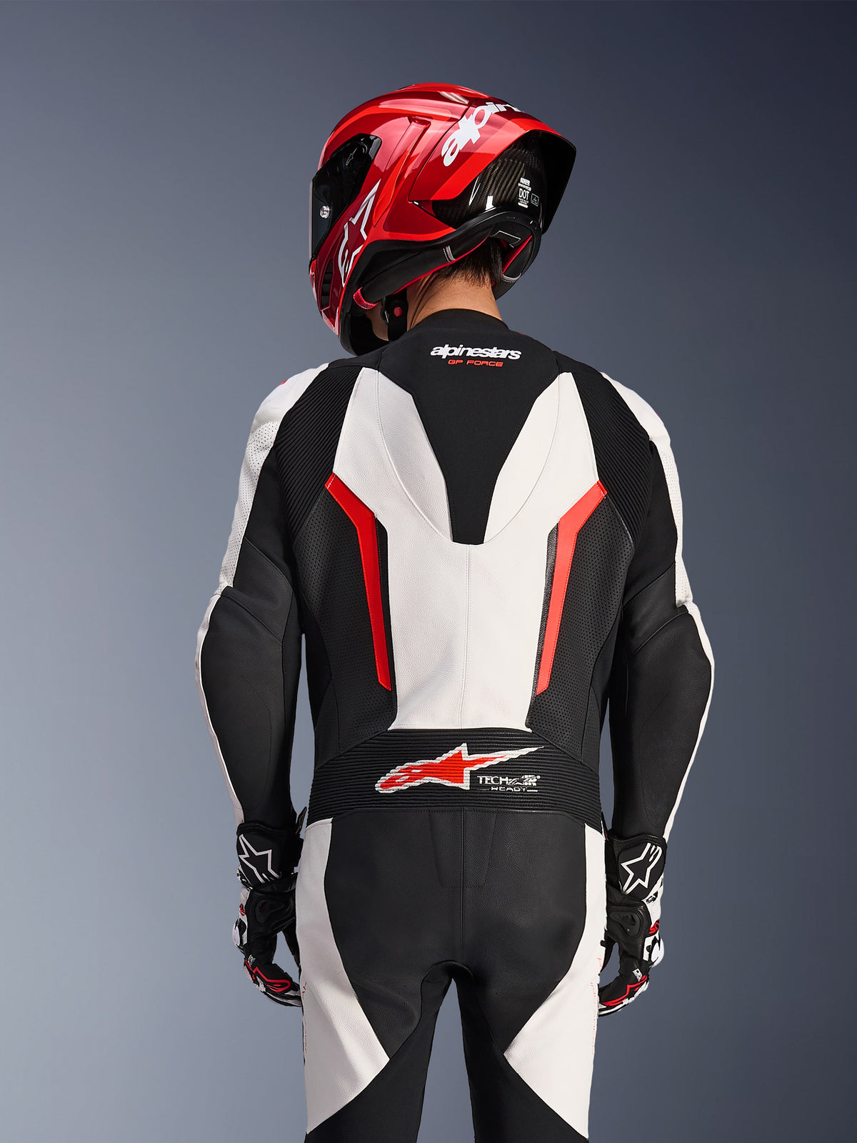 Vista traseira do fato de pele Alpinestars GP Force V2 de 1 peça em branco, preto e vermelho fluo, usado com um capacete de corrida vermelho, com a marca Tech-Air Ready na zona lombar, painéis de pele perfurada e zonas elásticas em sanfona para performance em corrida.