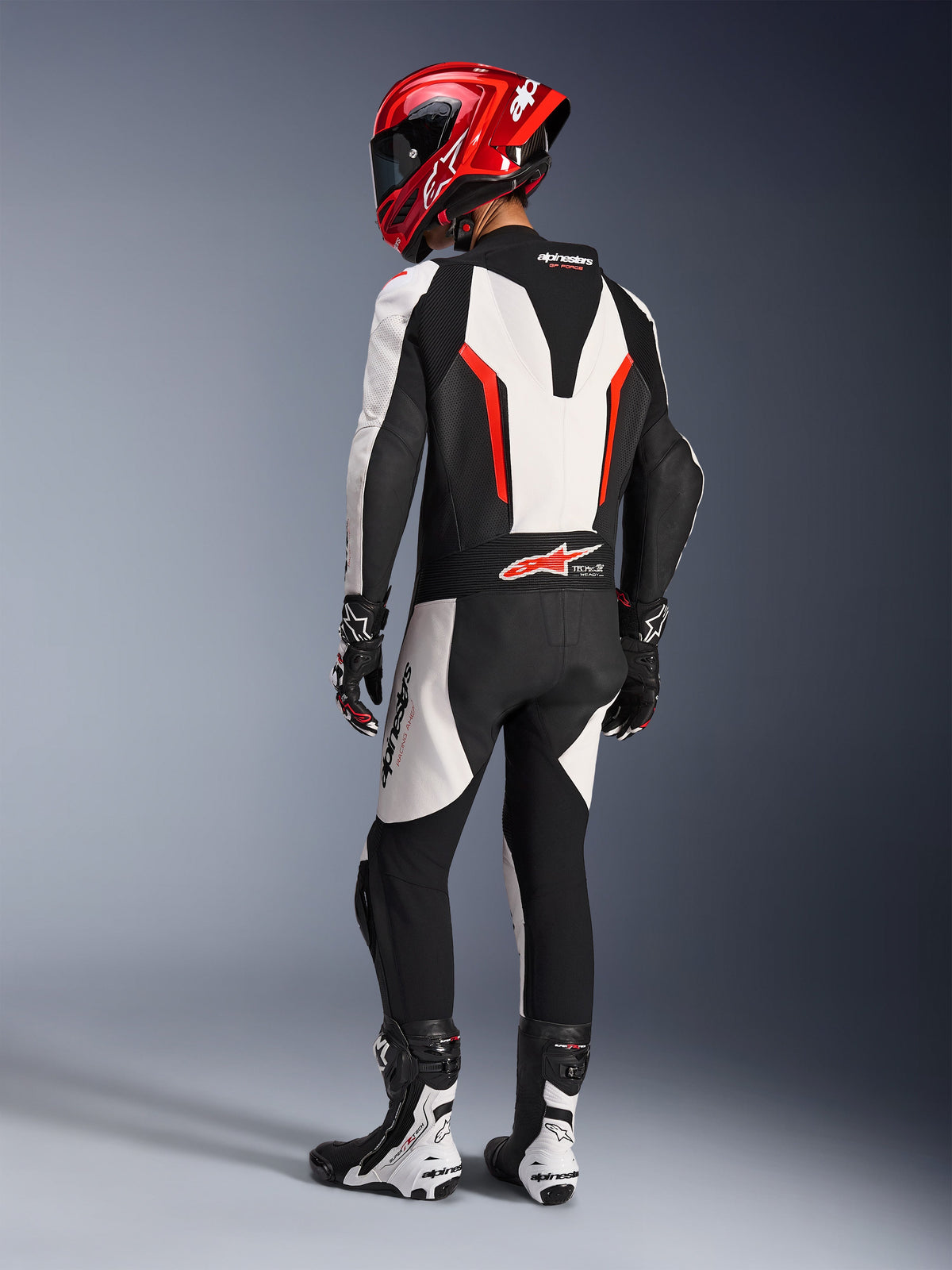 Alpinestars GP Force V2 Leather Suit - 1 Peça, Branco Preto Vermelho Fluo, vista traseira de piloto com equipamento completo de corrida, incluindo capacete vermelho, botas e luvas pretas e brancas, destacando a corcova aerodinâmica e os painéis elásticos ergonómicos.