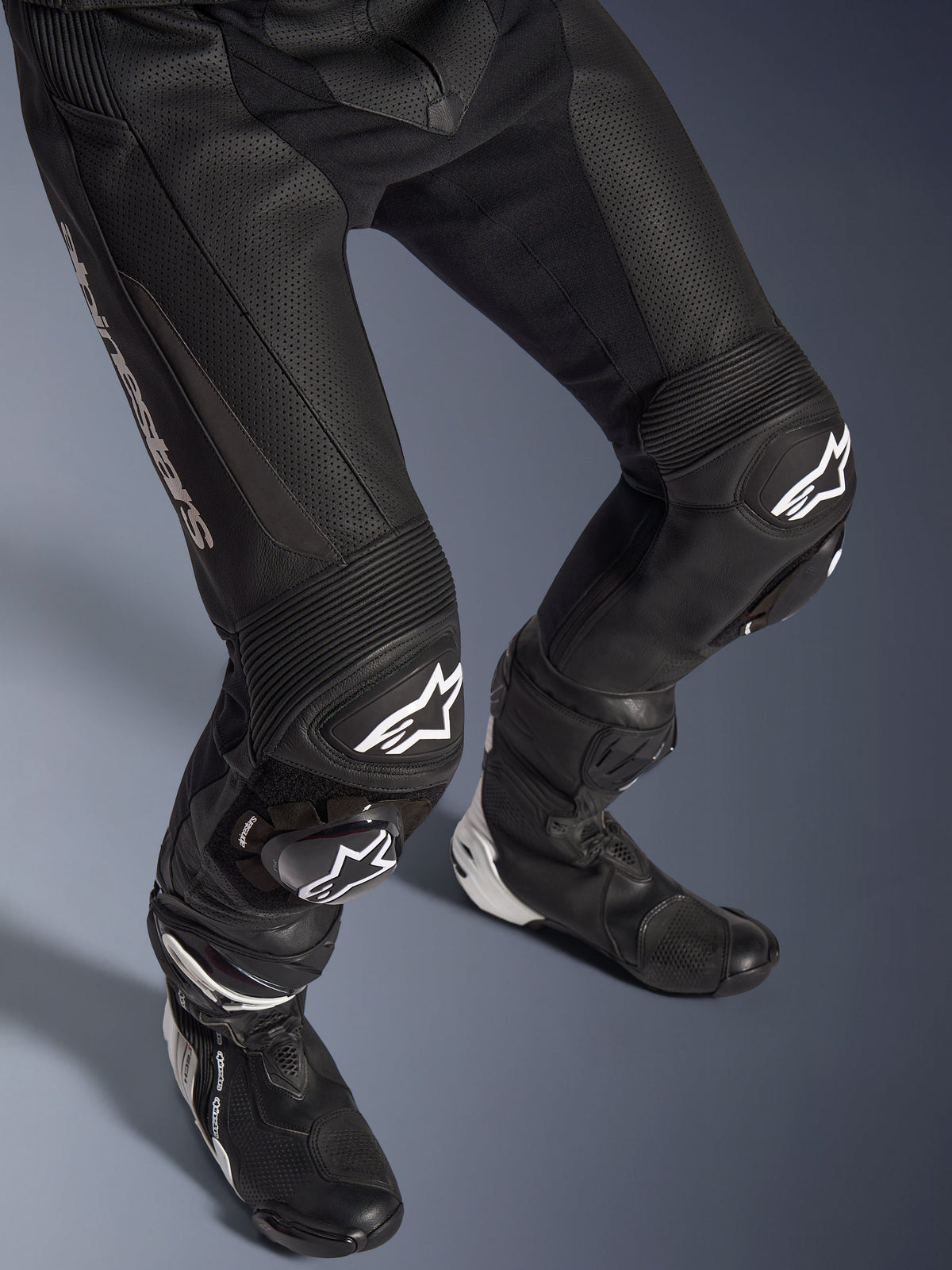 Alpinestars GP Force V2 2-Piece Leather Suit, close-up of the lower body including pants and racing boots, Black with white logos, featuring perforated leather construction, accordion stretch panels on the knees, and protective sliders, professional sport riding gear.